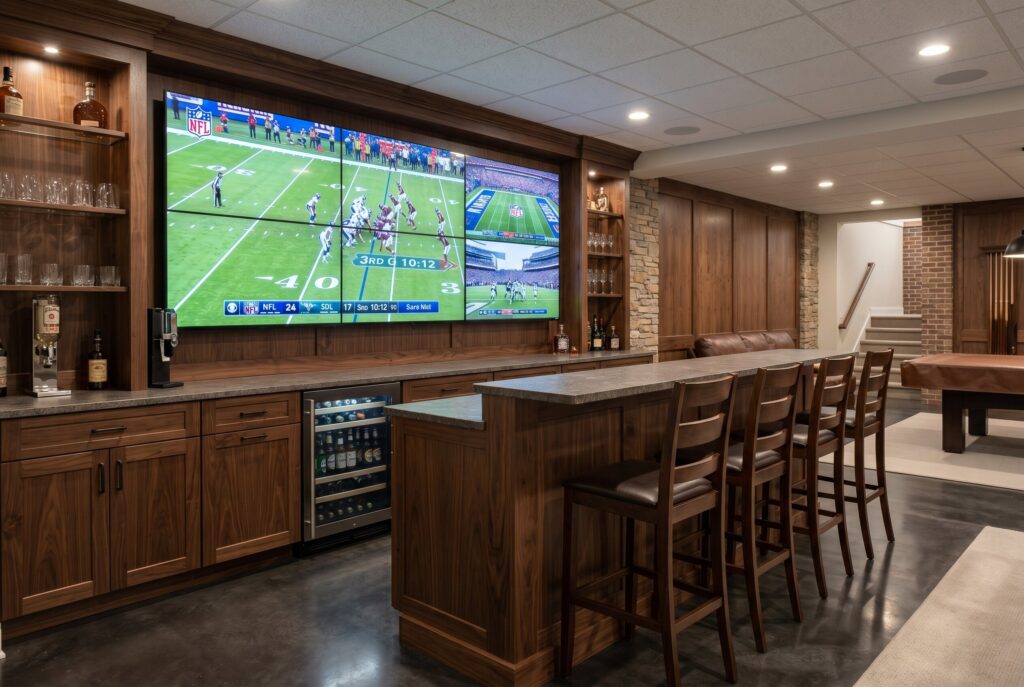 Multi screen walnut sports bar with video wall, stone countertop, and beverage storage.