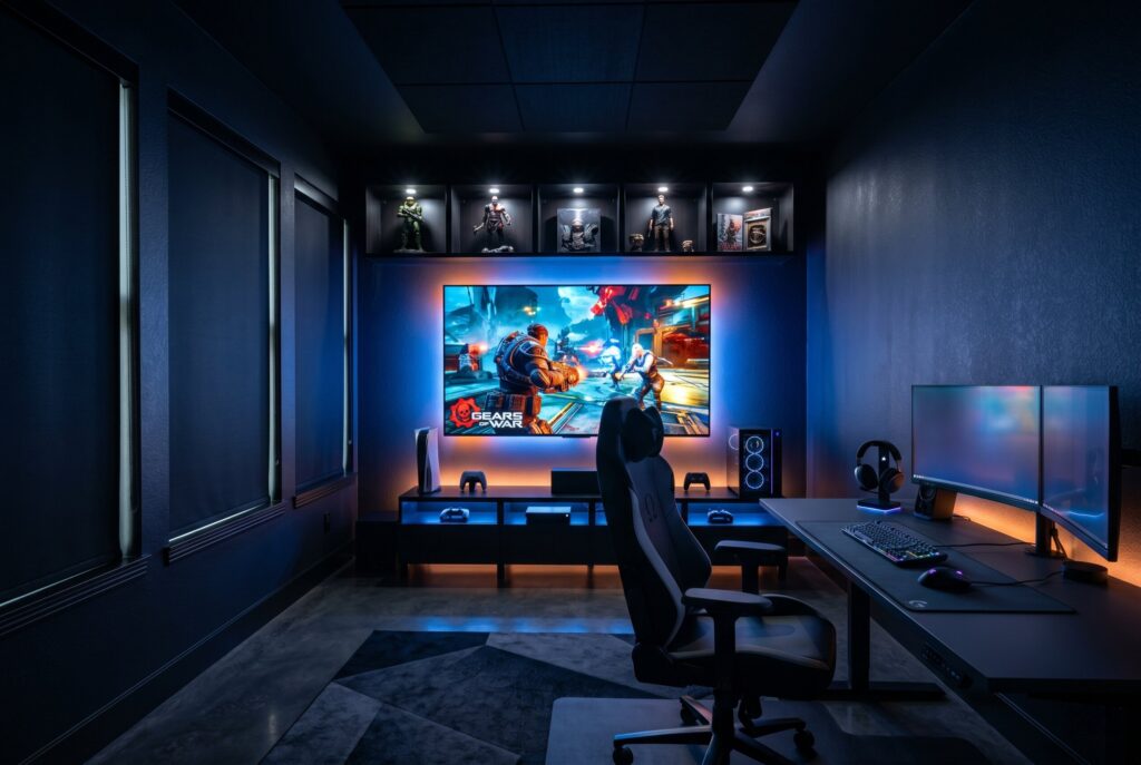 An immersive LED gaming cave painted matte black, featuring a desk with dual monitors, a large wall-mounted TV, indirect blue RGB lighting, and gaming collectibles on upper shelves.