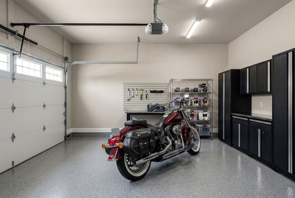 A brightly lit garage with a satin or semi-gloss finish on light walls, containing a red motorcycle, black cabinets, and a gray epoxy floor.