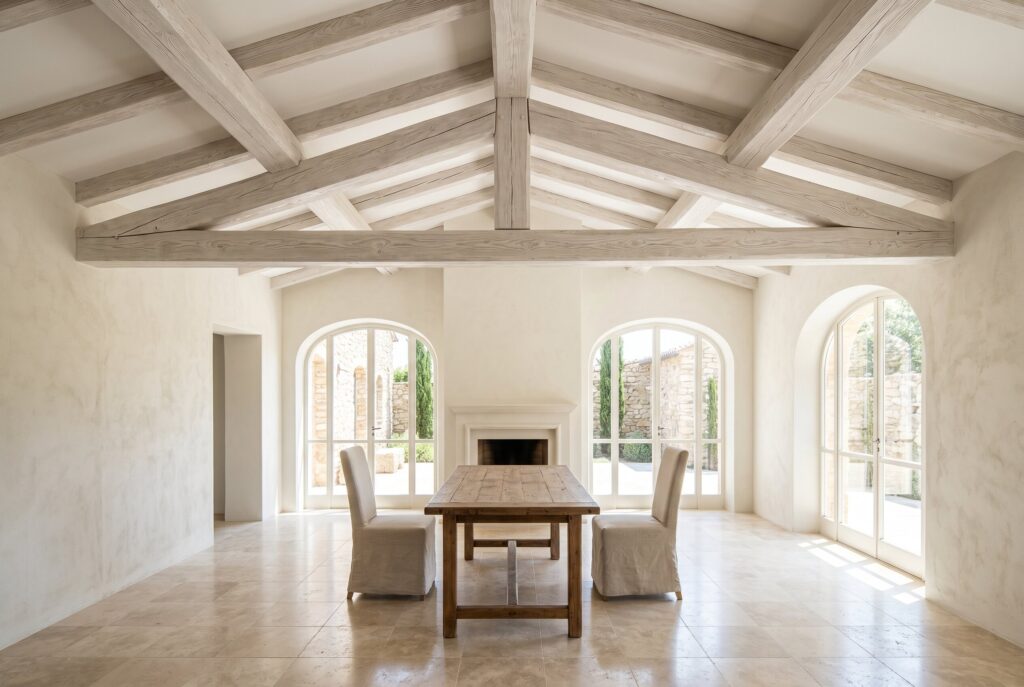 Bright airy room with whitewashed beams, warm white walls, a simple wooden table, two chairs, and arched glass doors.