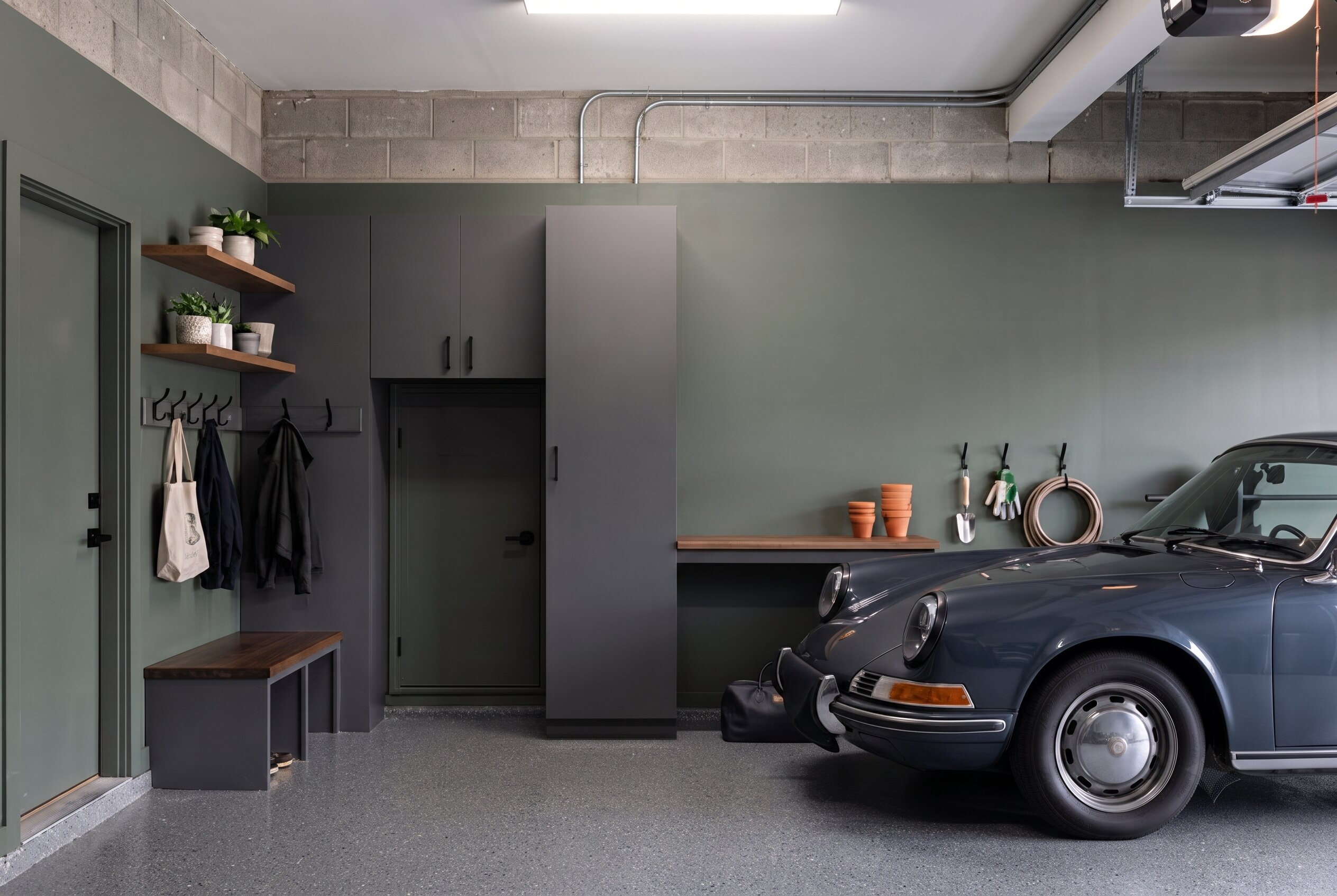 A garage doubling as a mudroom painted in Sherwin Williams Evergreen Fog, showing charcoal cabinets, a wooden bench, wall hooks, and a vintage blue Porsche.