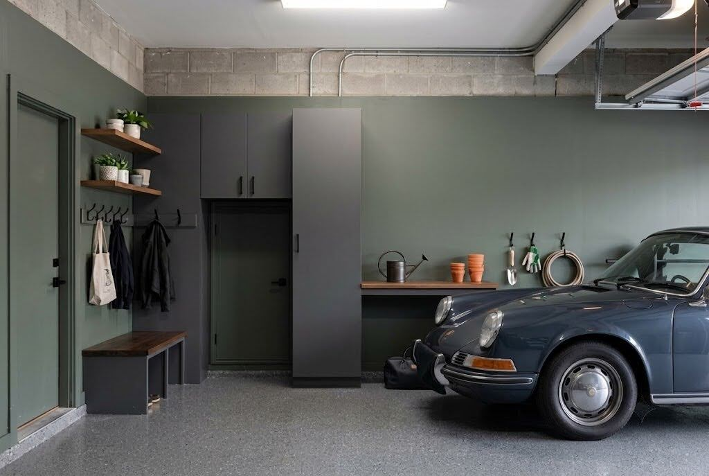 A garage doubling as a mudroom painted in Sherwin Williams Evergreen Fog, showing charcoal cabinets, a wooden bench, wall hooks, and a vintage blue Porsche.