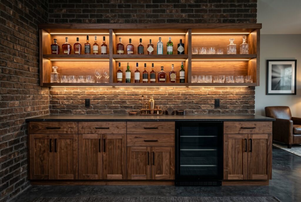 A one-wall dry bar featuring lower walnut cabinets, a mini-fridge, and upper glass shelves illuminated by warm LED strips holding various liquor bottles.