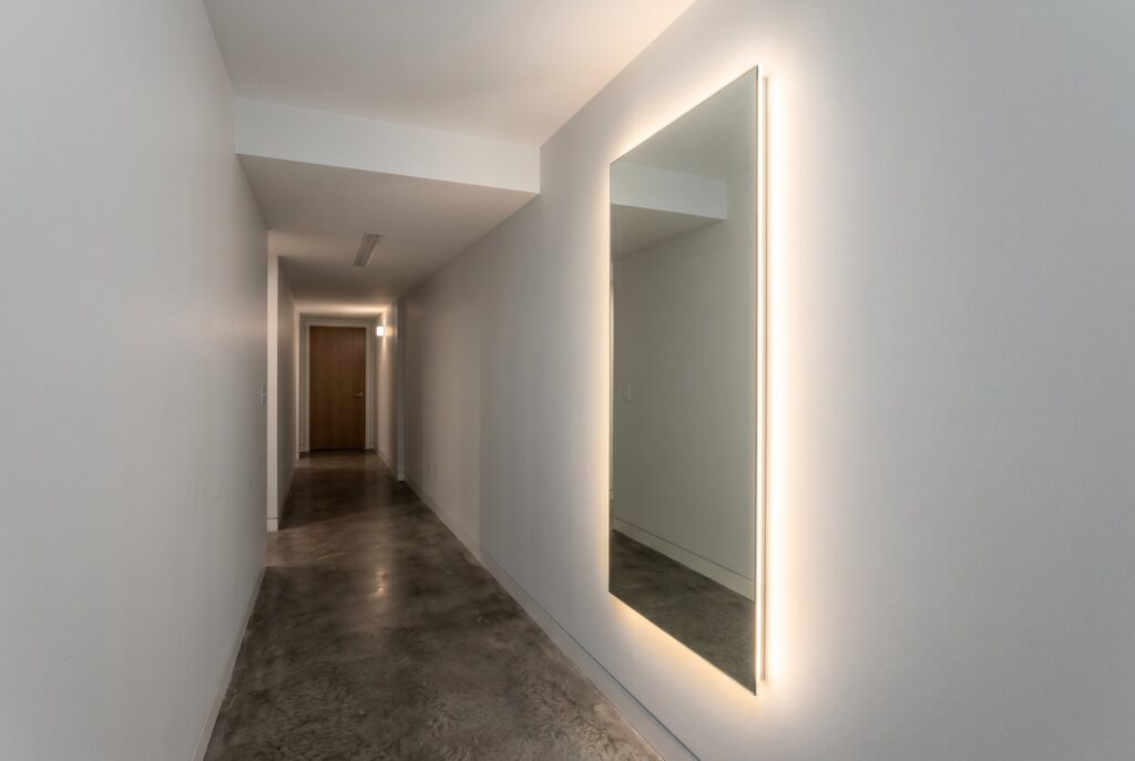 Tall backlit mirror expanding visual space in a modern basement hallway.