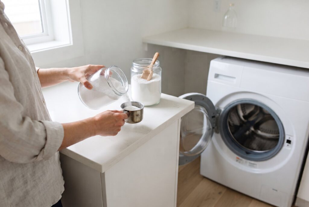 A person pouring white powder from a glass jar into a metal measuring cup in a laundry room. 