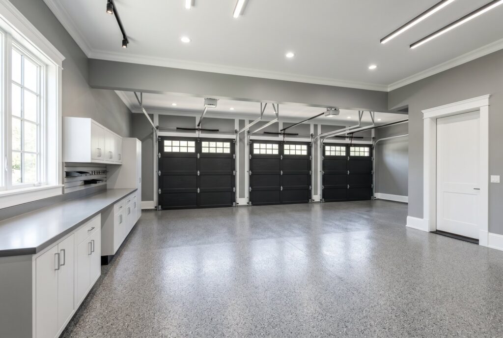 A large garage painted in Benjamin Moore Coventry Gray with three black garage doors, white trim, white cabinets, and gray flake epoxy floors.
