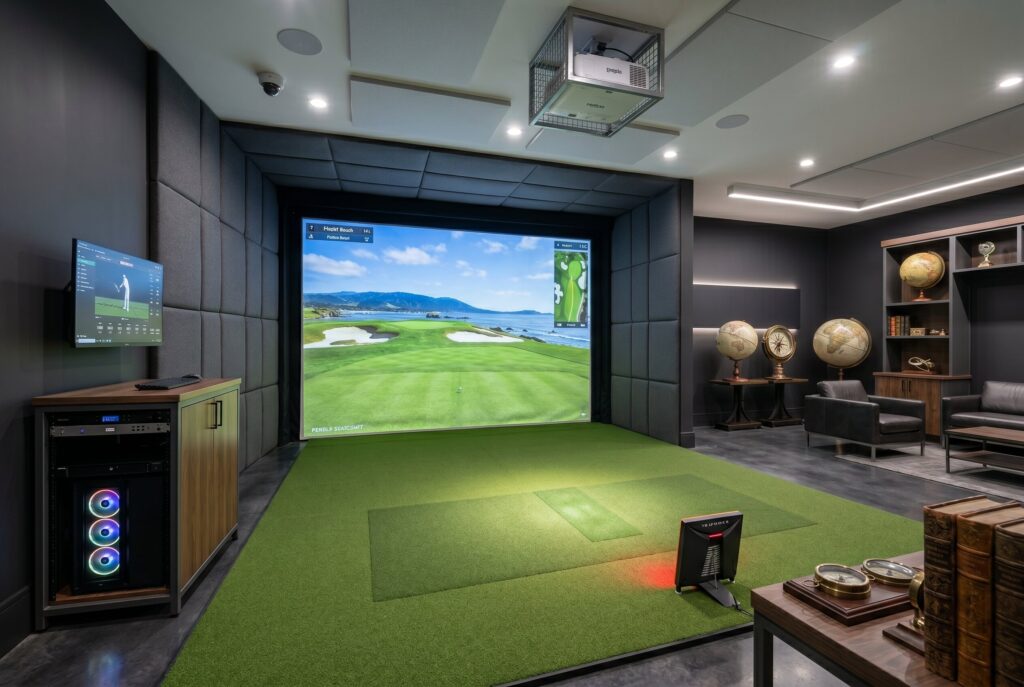 An indoor golf simulator room with green turf, padded dark walls, a large screen displaying a golf course, and a seating area with leather chairs and globes on a console.