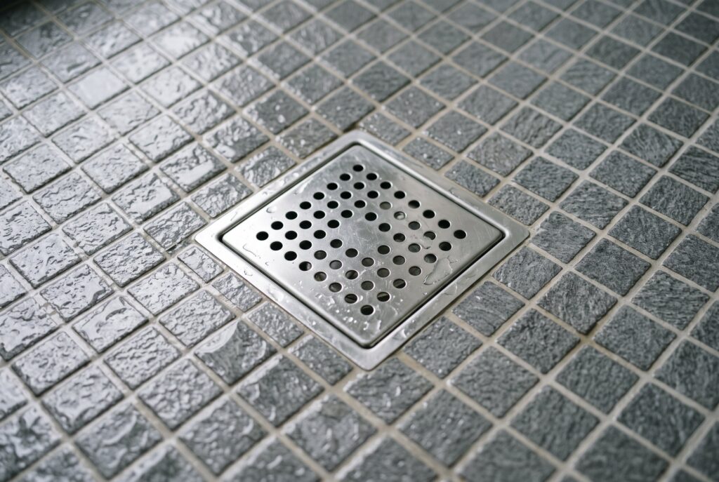 Square metal shower drain surrounded by wet gray mosaic tiles.