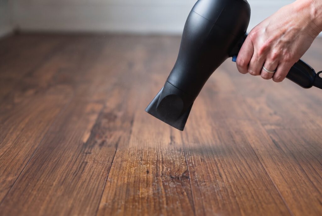 A hand using a hairdryer to soften old, hardened wax on a hardwood floor.