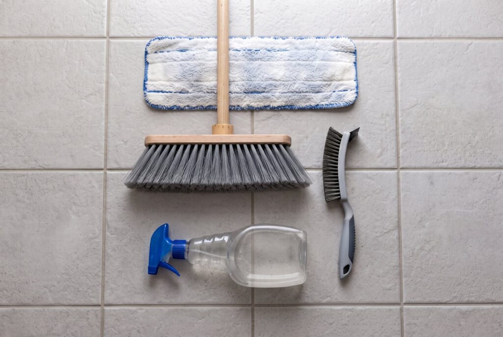 A flat-lay of cleaning tools including a mop pad, broom, grout brush, and spray bottle on a tiled floor.