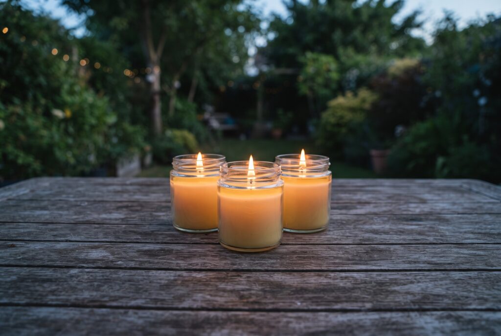 Three small glass jars with burning candles placed together on a rustic wooden table outdoors.
