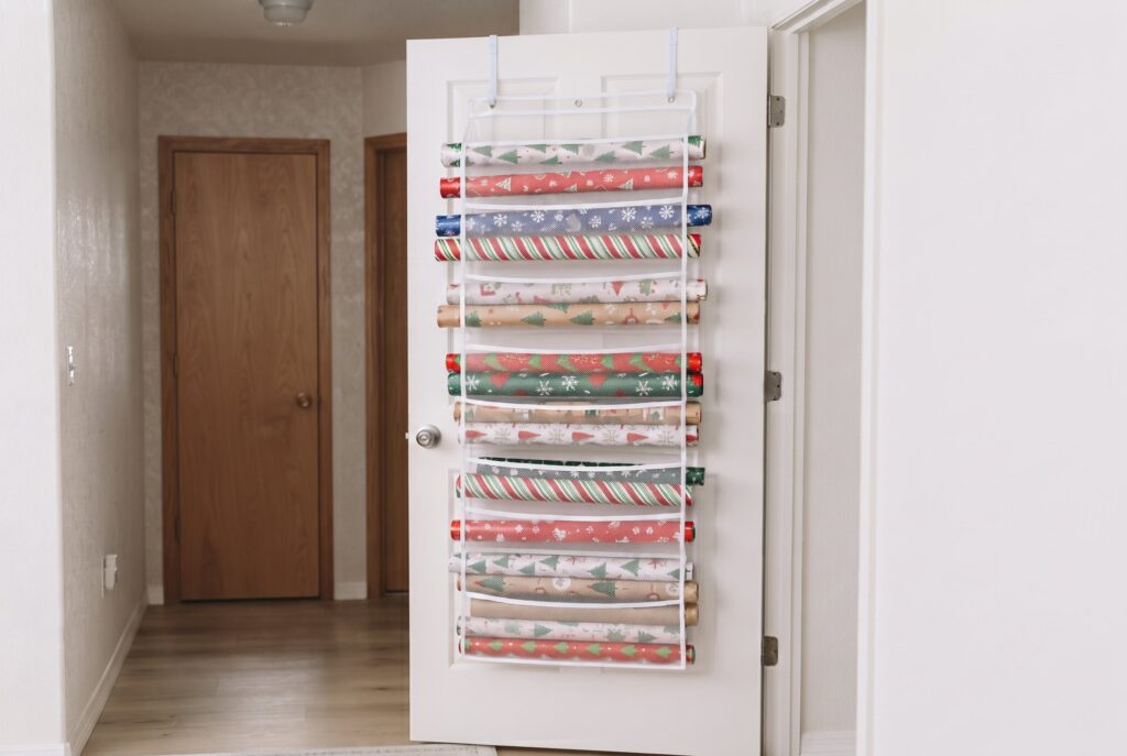A vertical white rack hanging over a door, filled with various rolls of holiday wrapping paper.