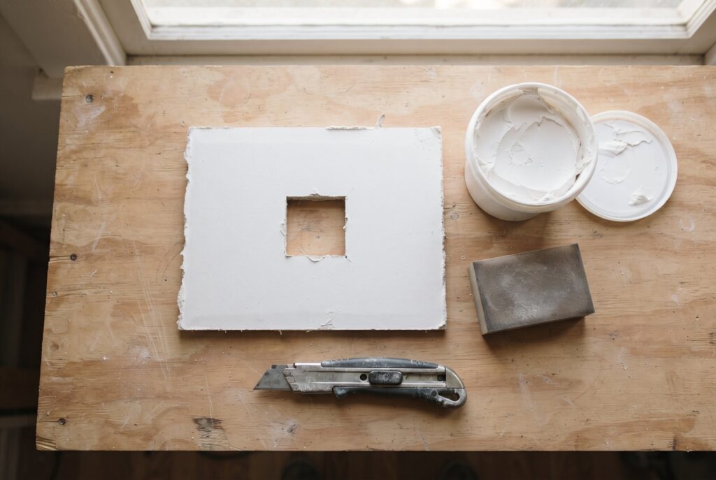 A piece of drywall, a utility knife, joint compound, and a sanding block on a wooden surface.