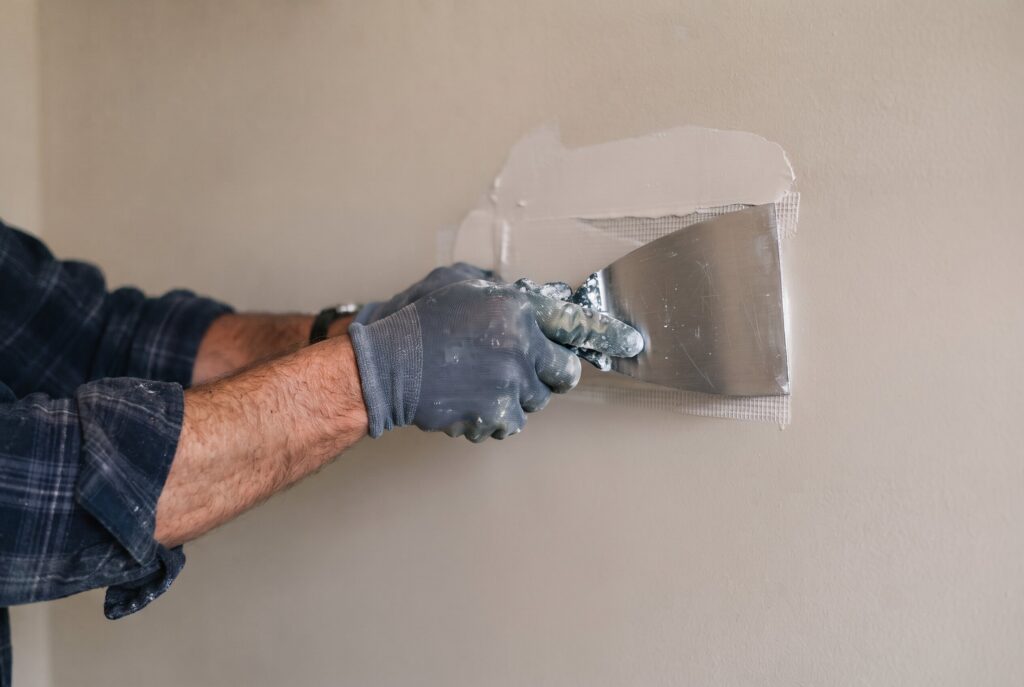 A person using a putty knife to spread joint compound over a repaired drywall patch.