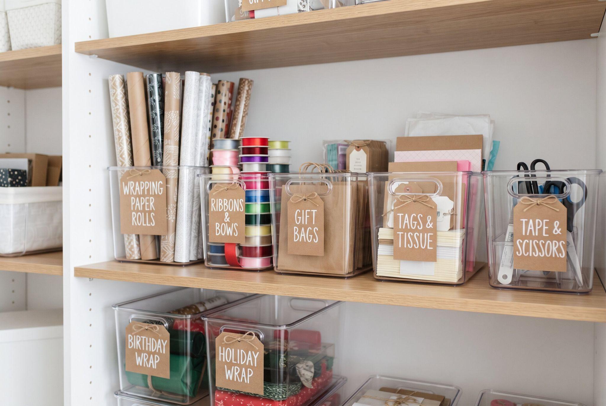 Clear storage bins on a shelf, each labeled with cardstock tags for different wrapping supplies.