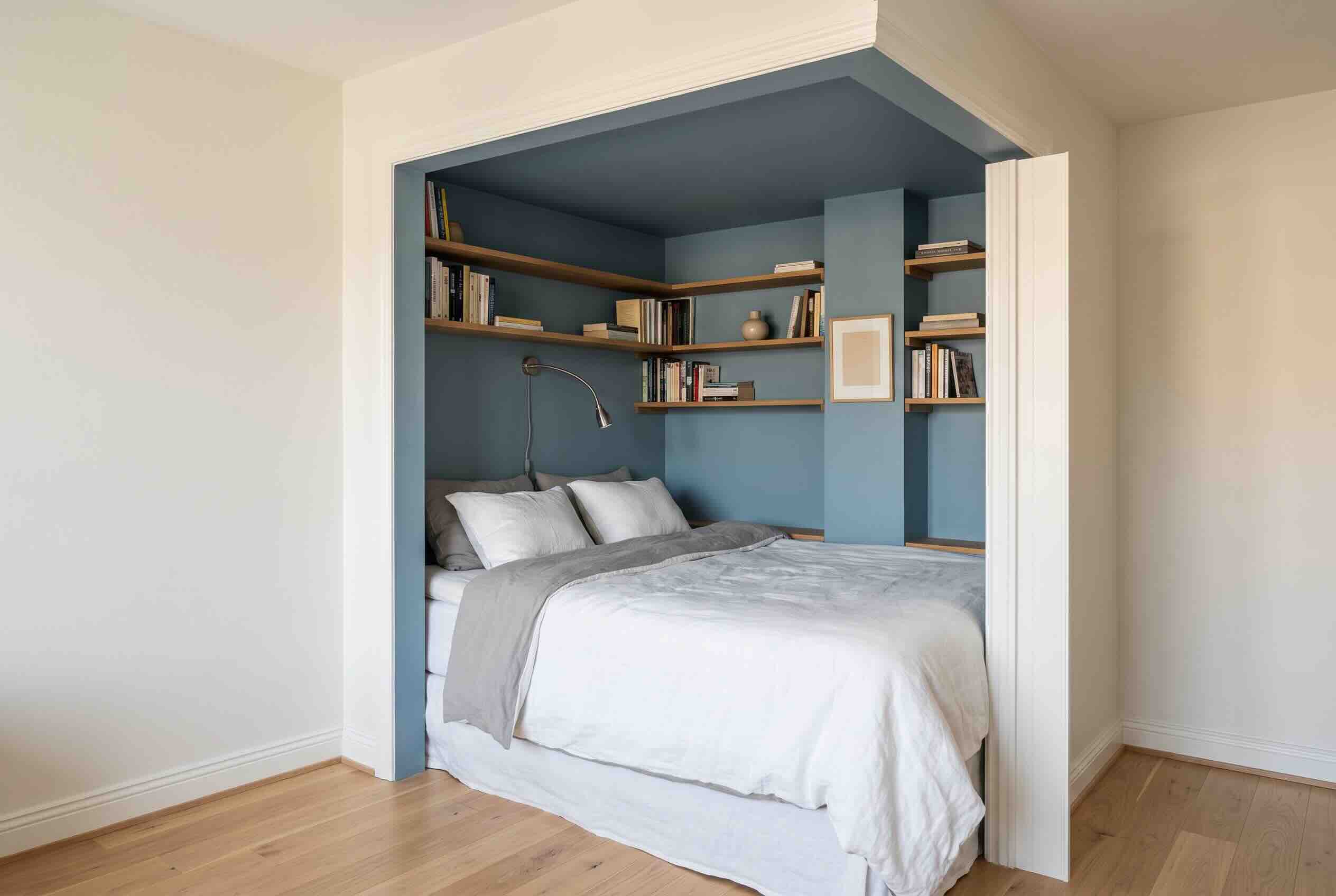 A bed tucked into a built-in framed alcove with blue walls and floating shelves.