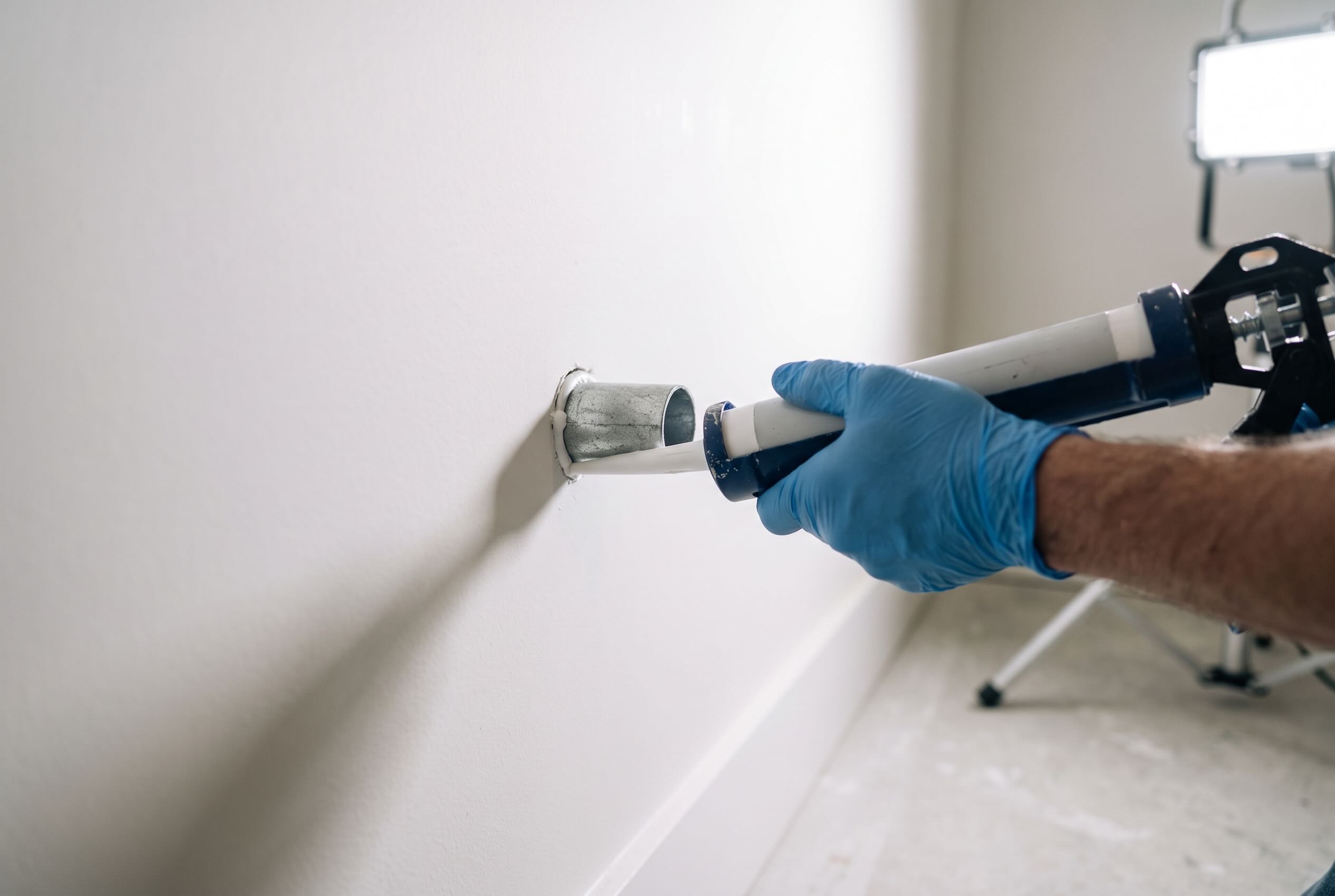 A caulking gun applying sealant around a metal pipe penetrating a white interior wall.