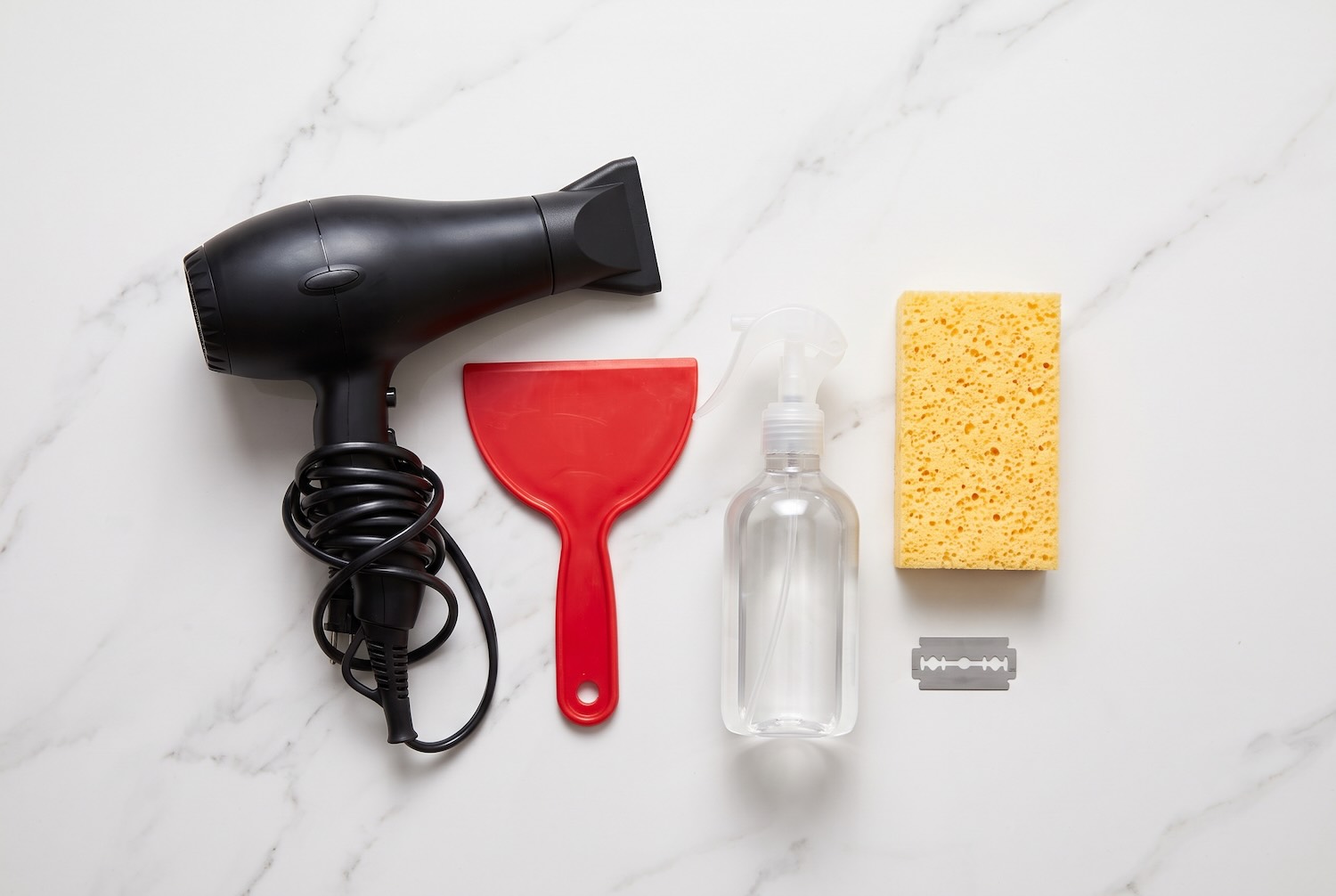 A collection of wallpaper removal tools including a hairdryer, plastic scraper, spray bottle, sponge, and razor blade.