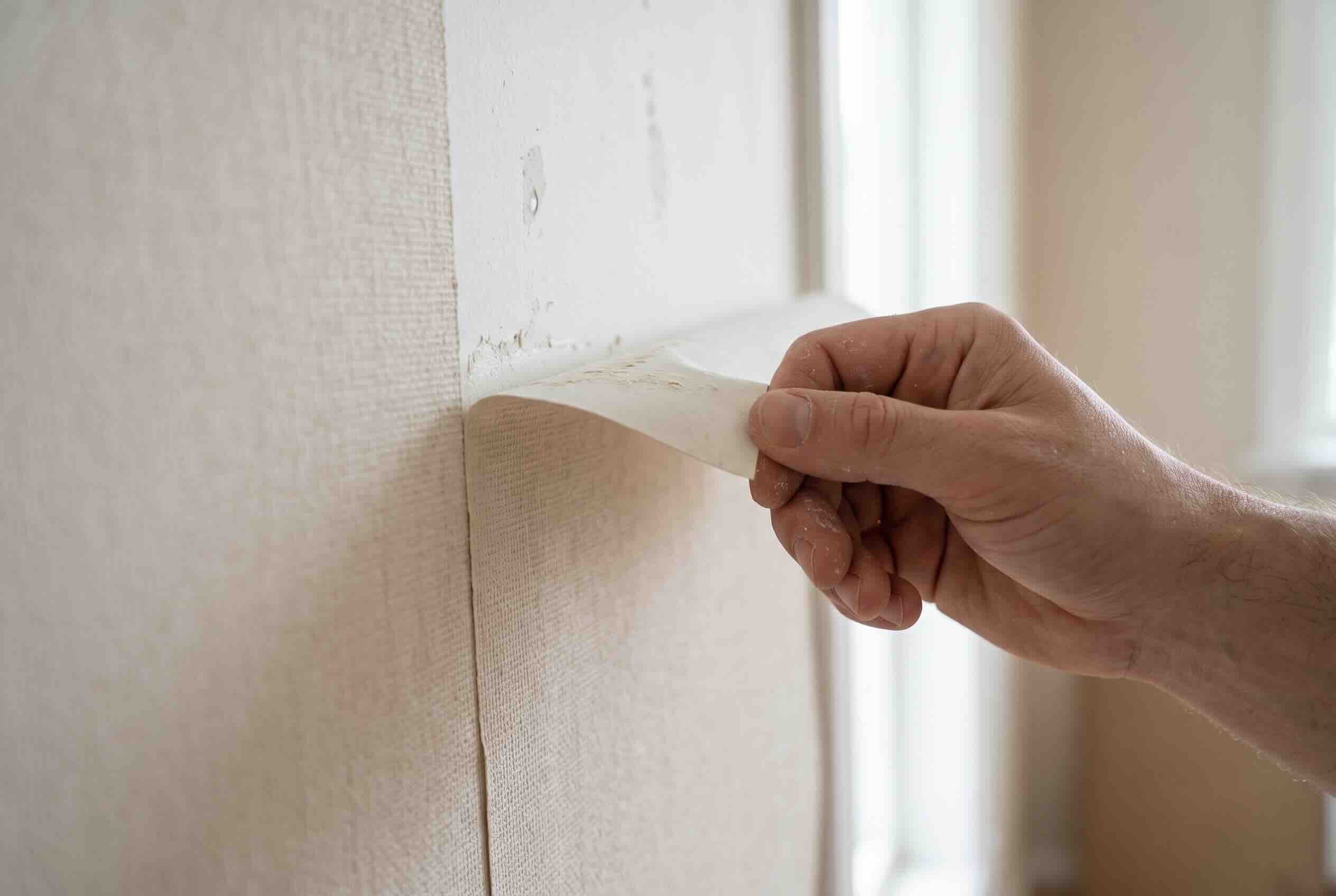 A hand lifting and peeling back the top corner of a strip of wallpaper.