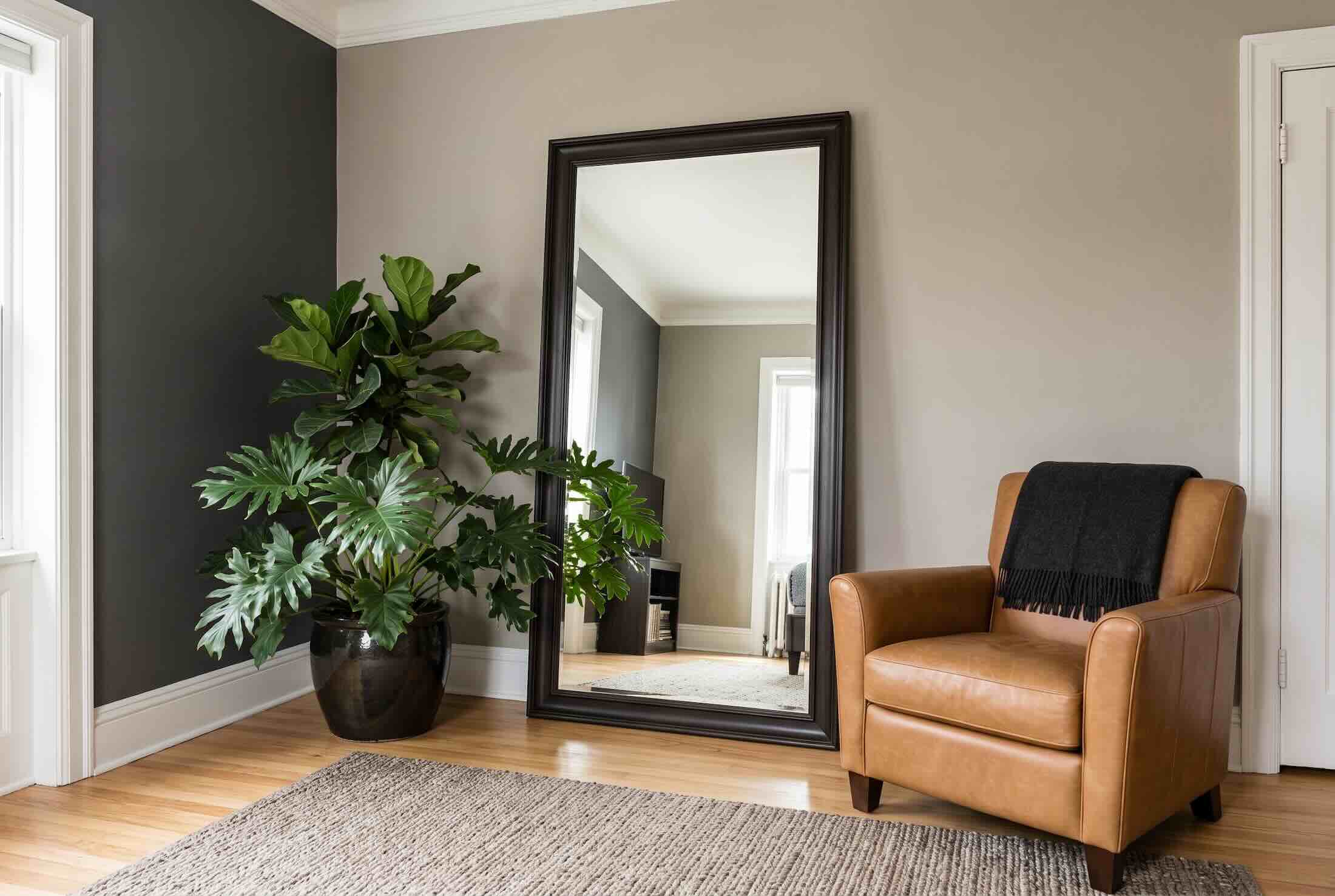 A large mirror leaning against a dark accent wall next to a leather armchair and a potted plant.