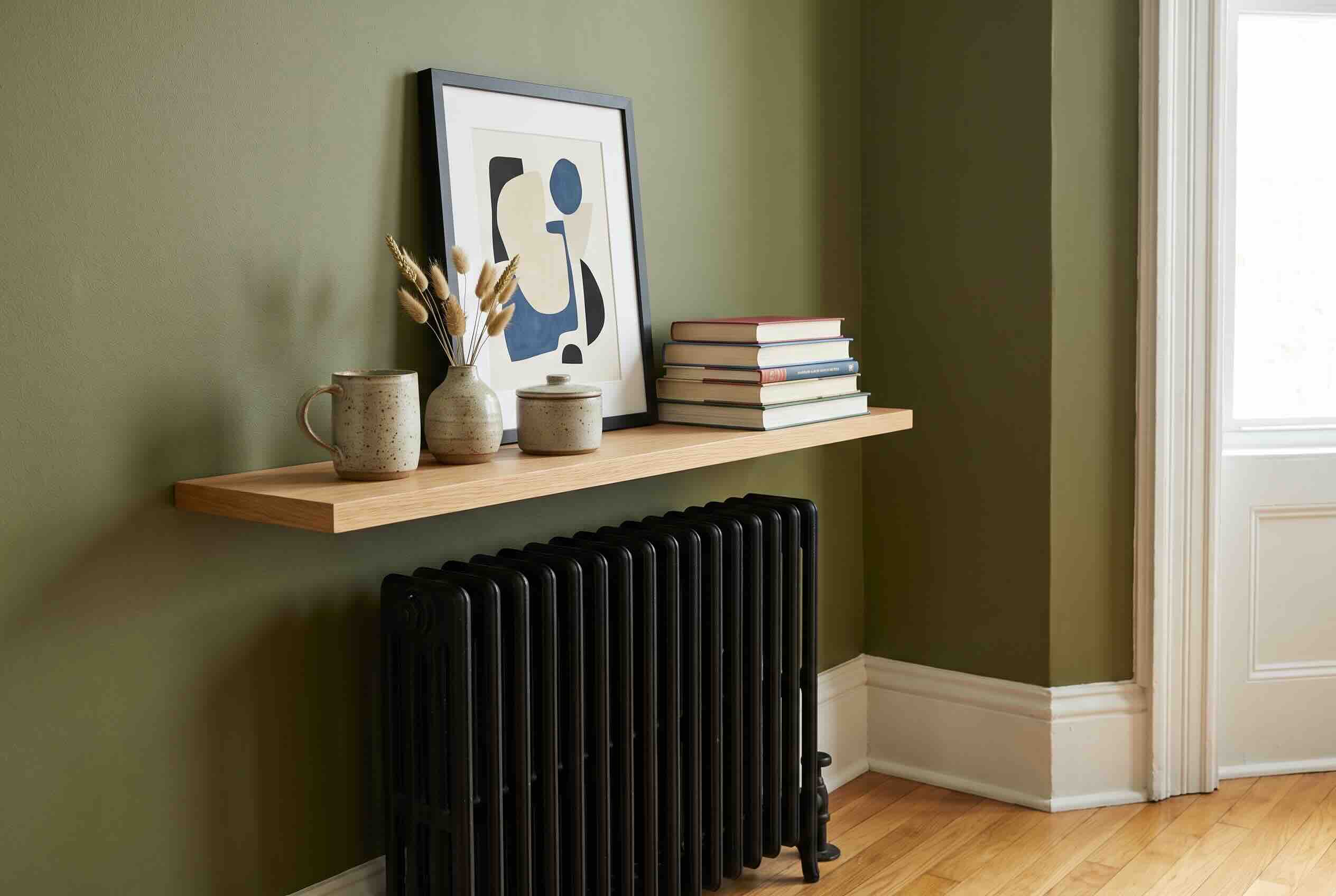 A light wood floating shelf mounted on a green wall directly above a black radiator.