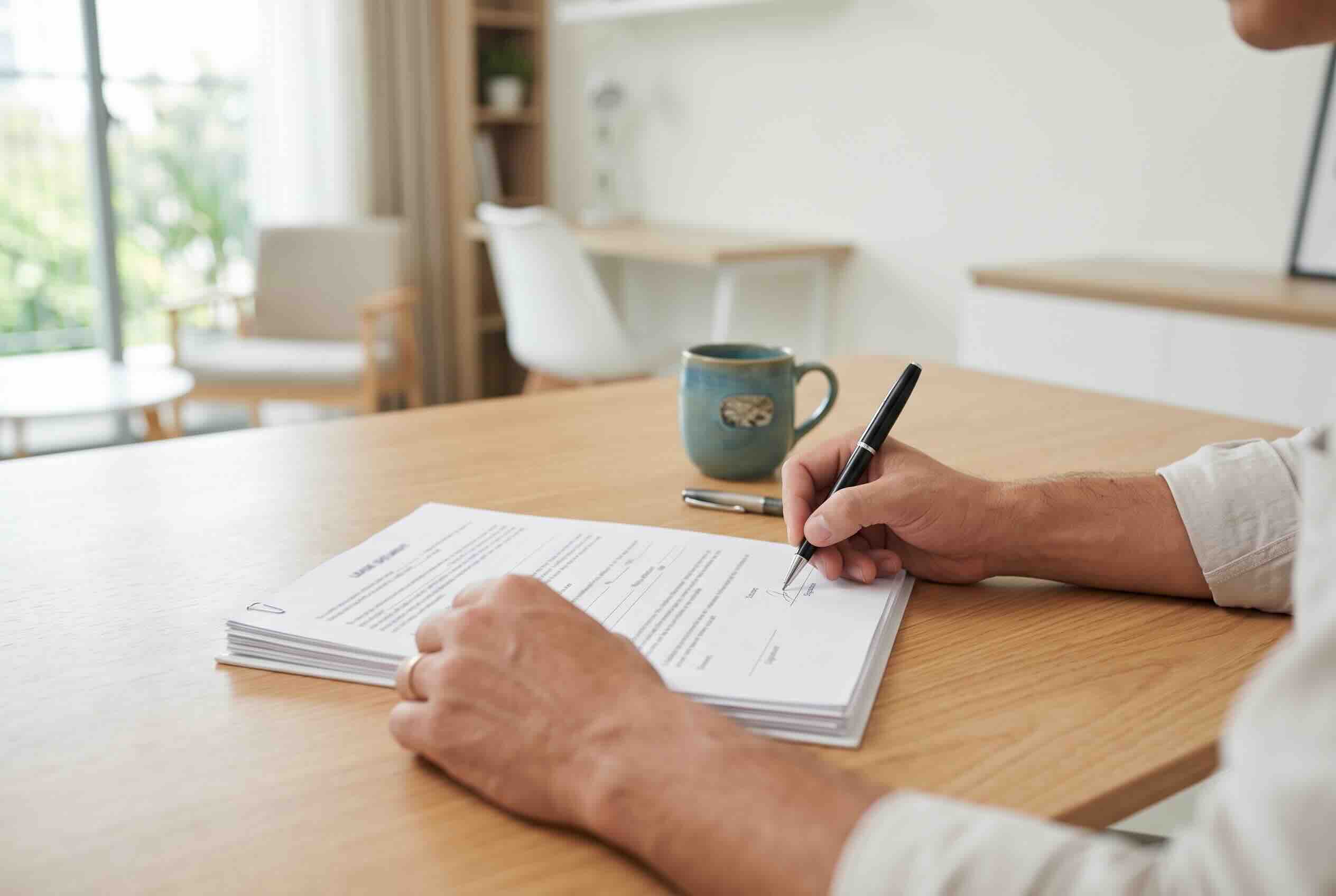 A person signing a document on a wooden desk with a coffee mug nearby.