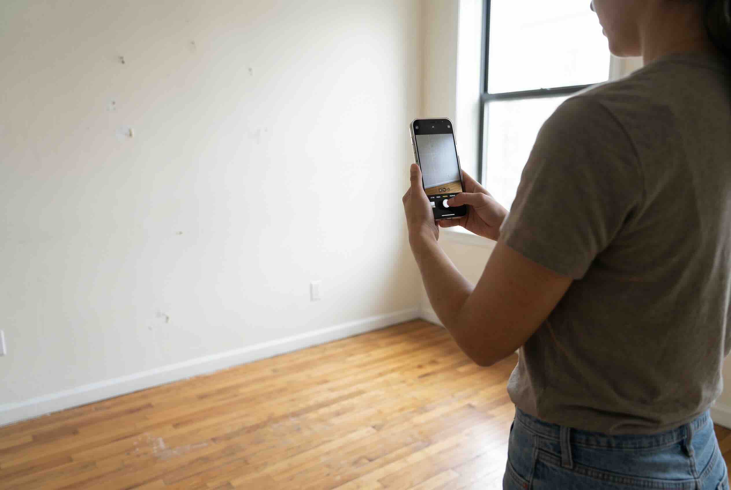 A person taking photographs of an empty apartment room with a smartphone.