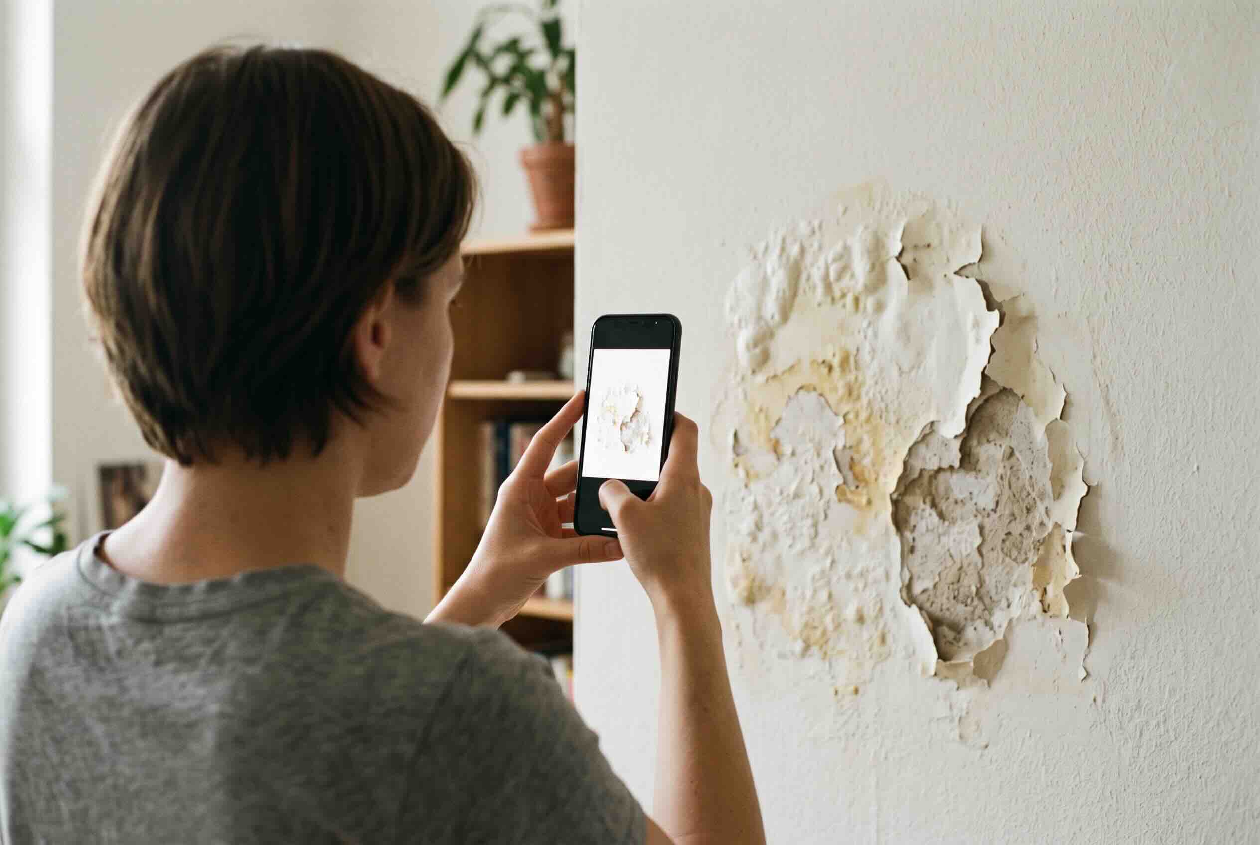 A person using a smartphone to document peeling paint and water damage on a wall.