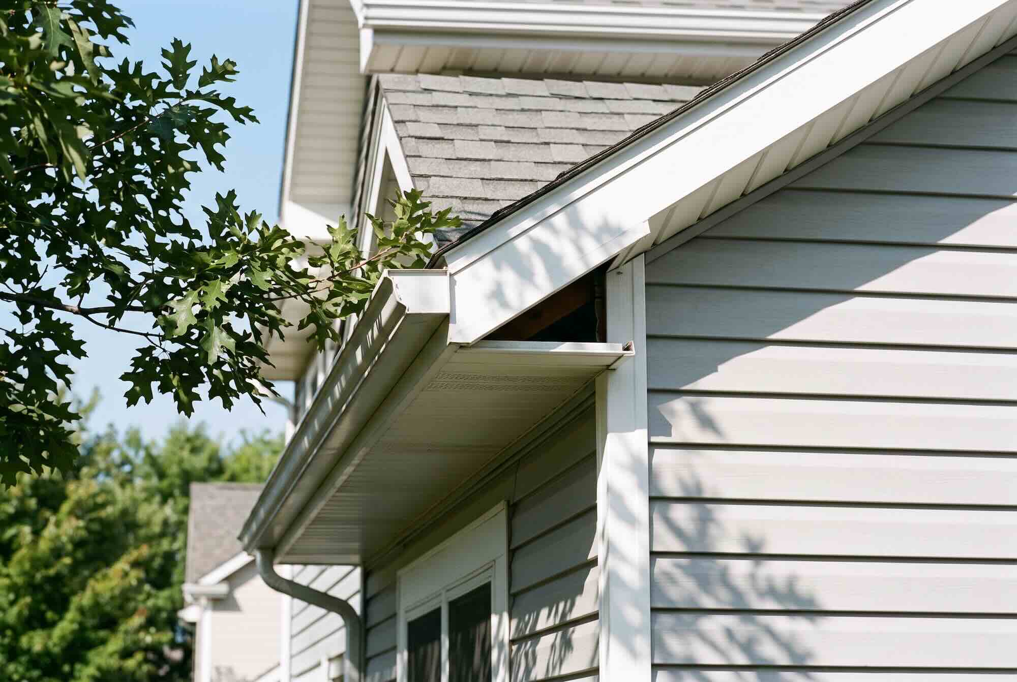 An unsealed gap between white roof fascia and grey siding near a house gutter.