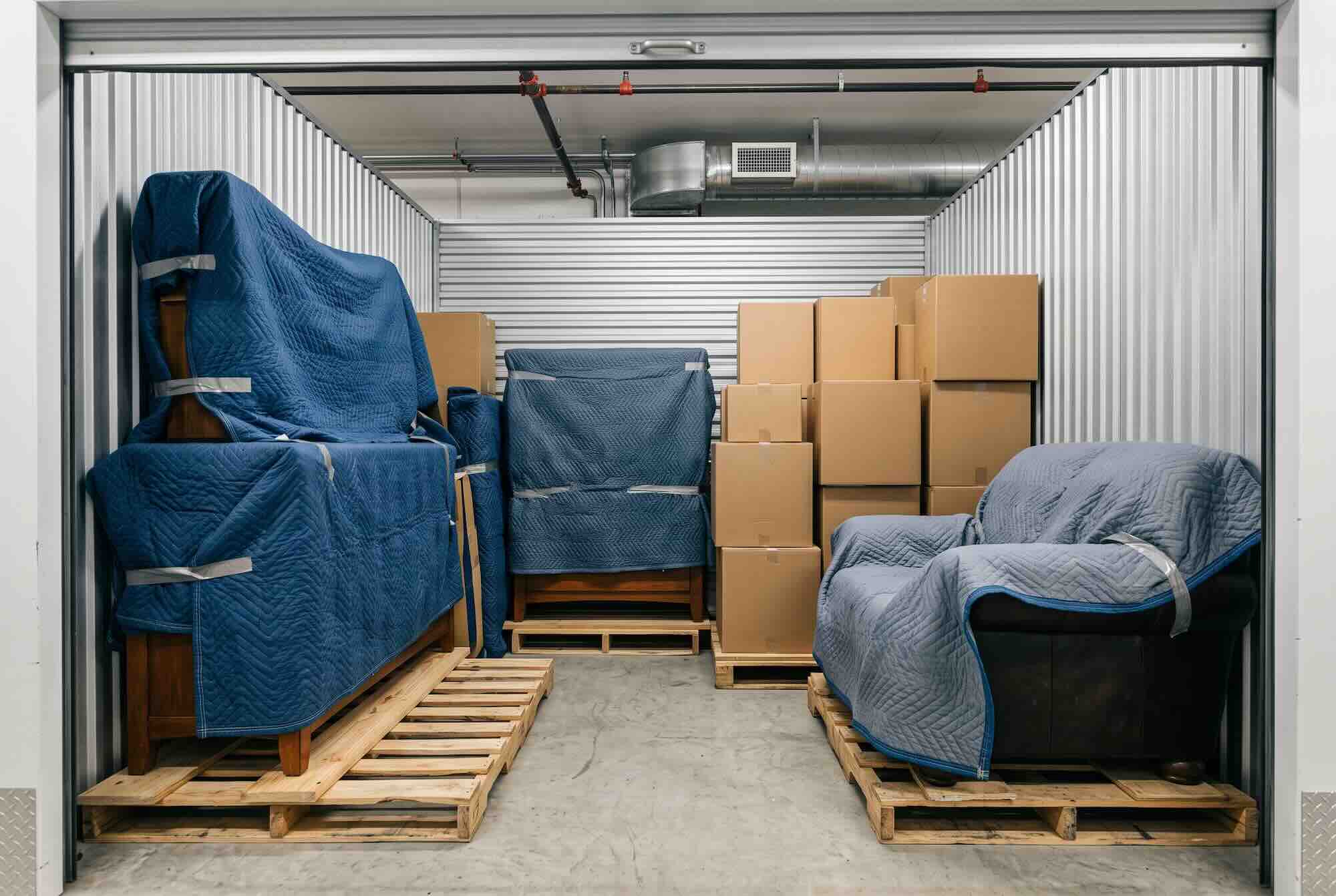 Blanket-wrapped furniture on pallets and stacked boxes in a storage unit.