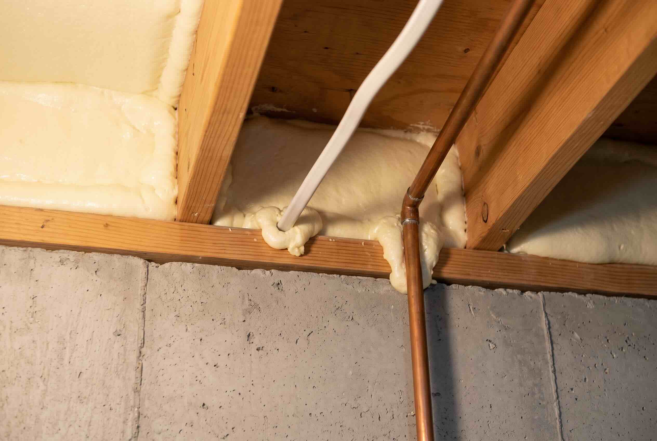 Cured closed-cell spray foam sealing the gaps around an electrical cable and a copper water pipe at a basement rim joist.