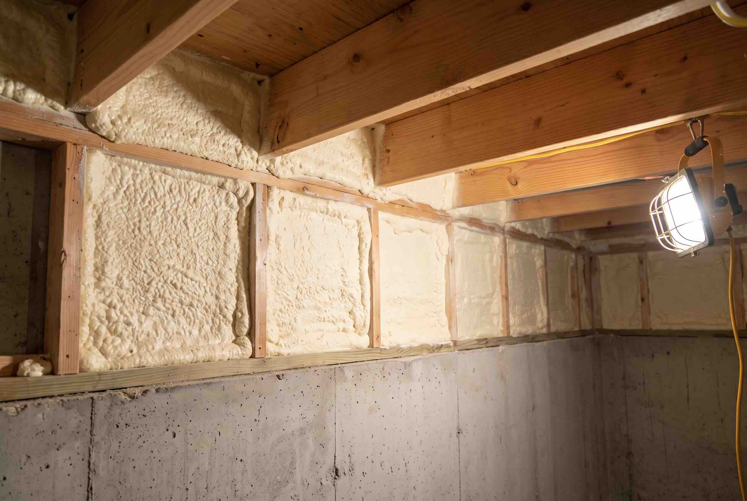 Cured pale yellow closed-cell spray foam expanding between wooden wall studs and floor joists in a basement.