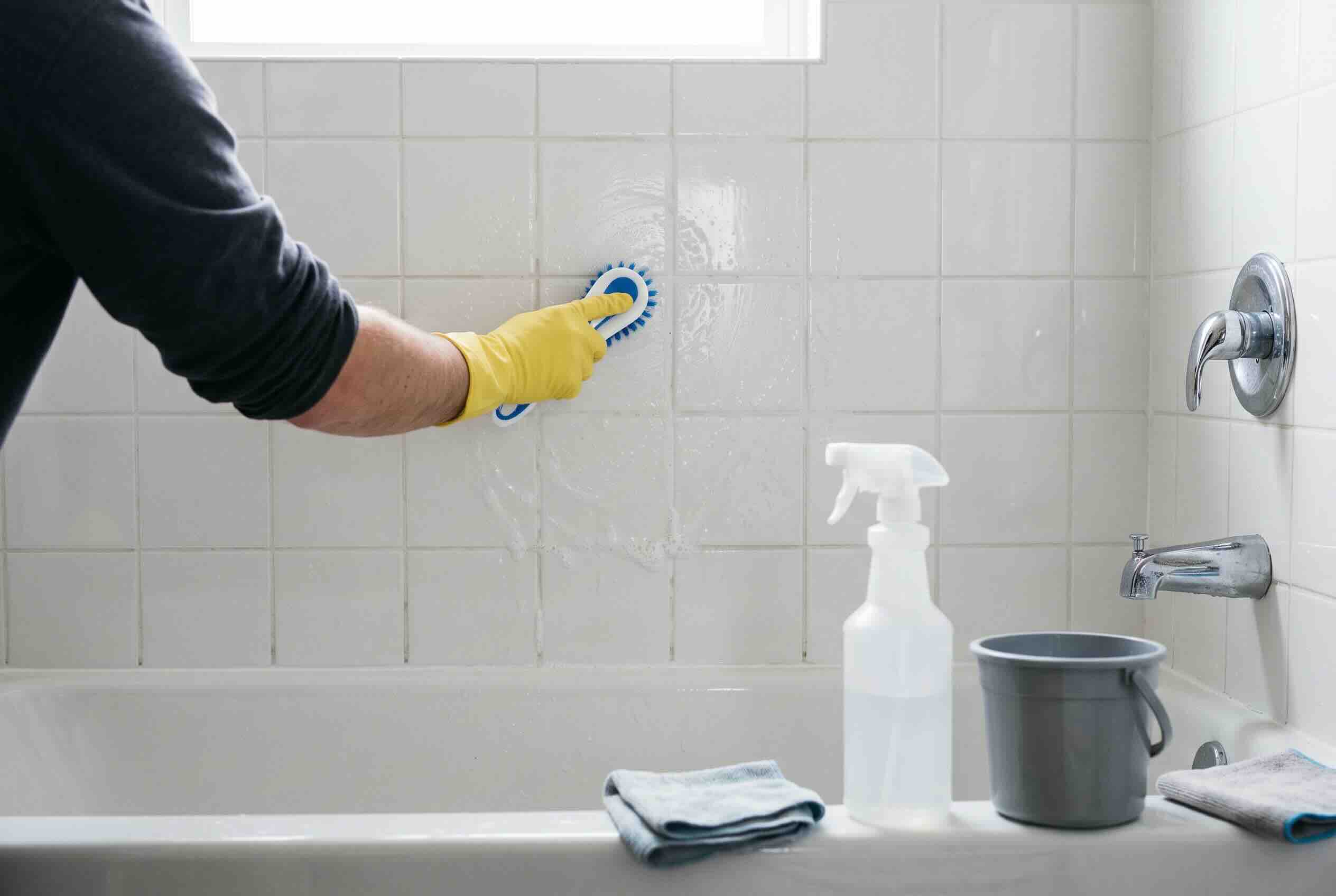 Scrubbing white bathroom wall tiles above a bathtub using a handled brush and cleaning spray.