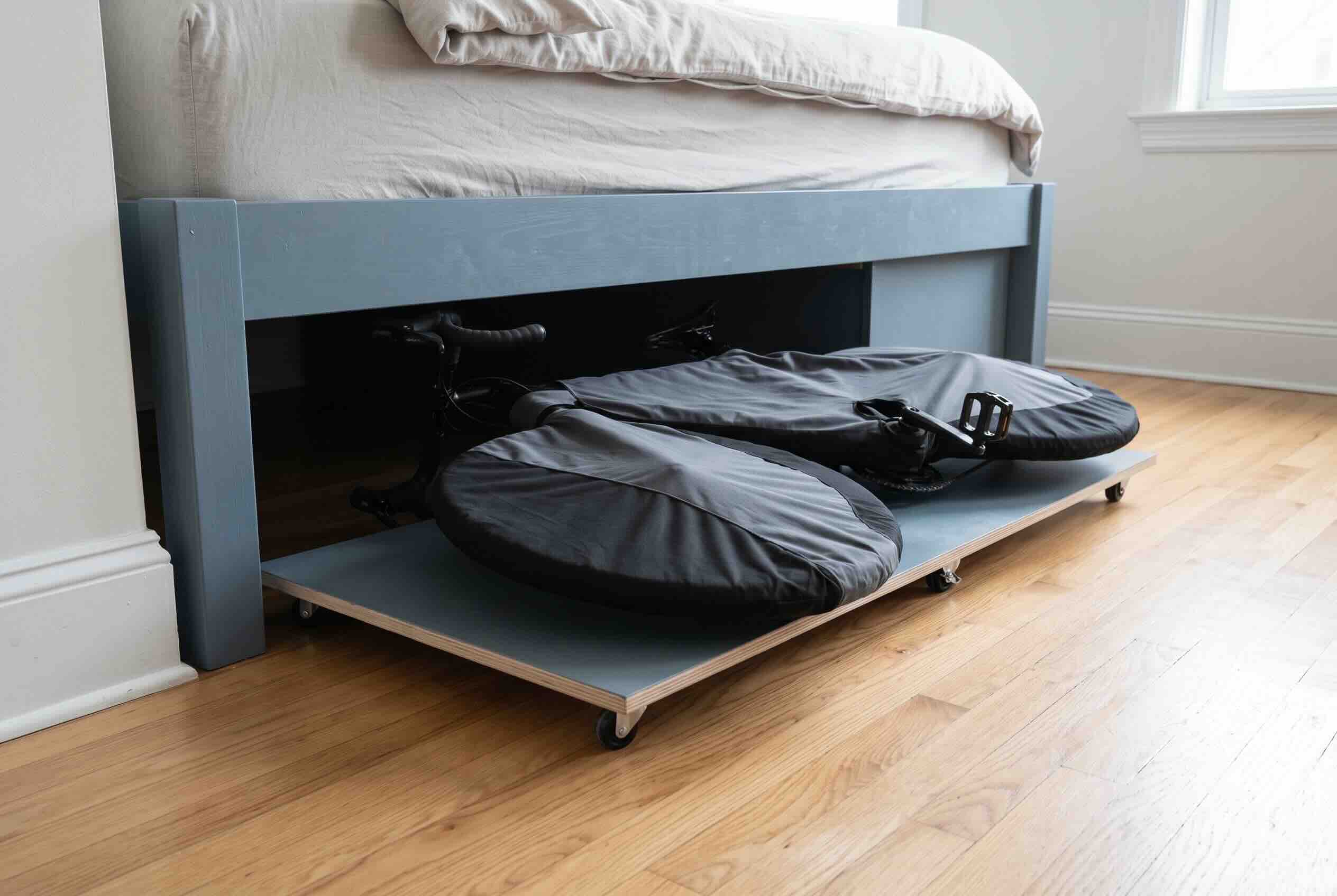 Sliding bicycle covered in black protective bags under a bed using a rolling wooden tray.