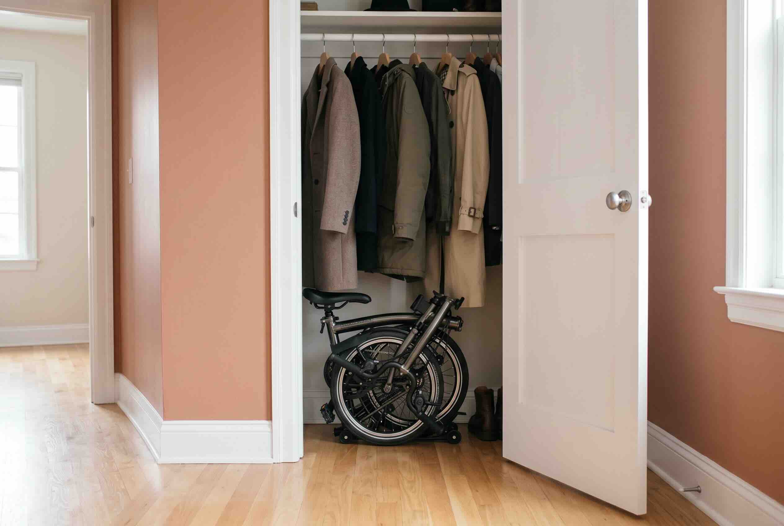Storing a folded black bicycle on the floor inside a hallway closet beneath hanging coats.