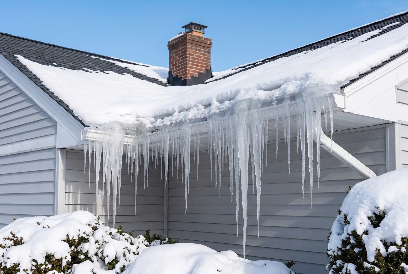 Thick icicles hanging from a snow-covered roof and a clogged gutter, forming an ice dam.