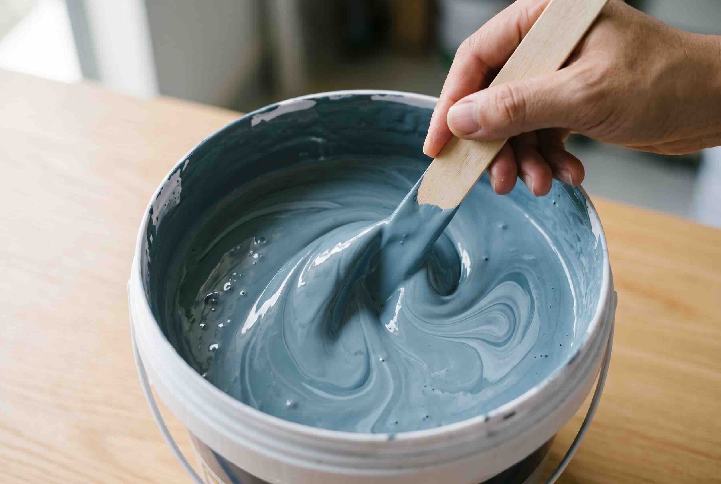 Thoroughly mixing bluish-grey paint in a bucket with a wooden stir stick.