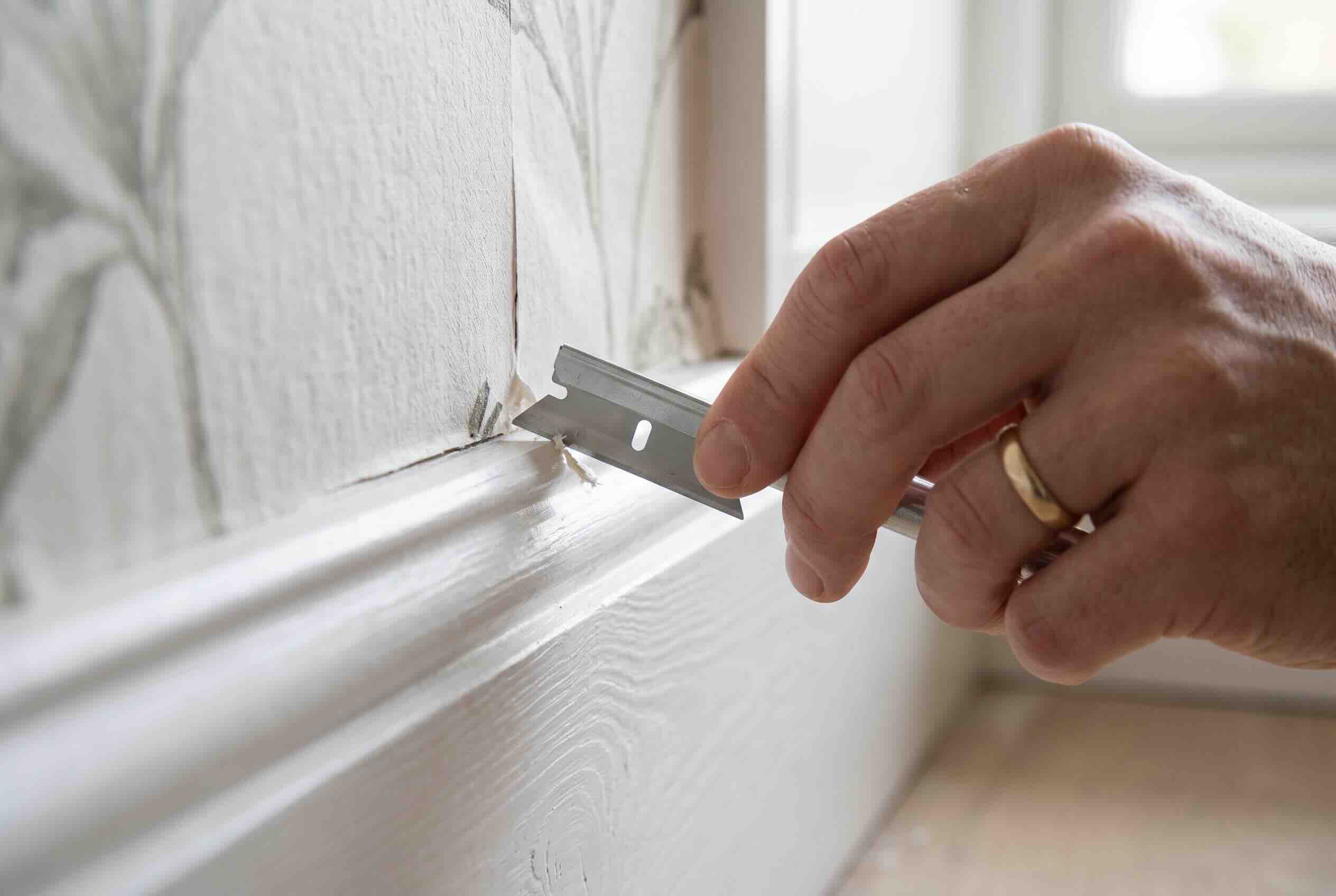 Using a razor blade to carefully score and free wallpaper trapped behind baseboard trim.