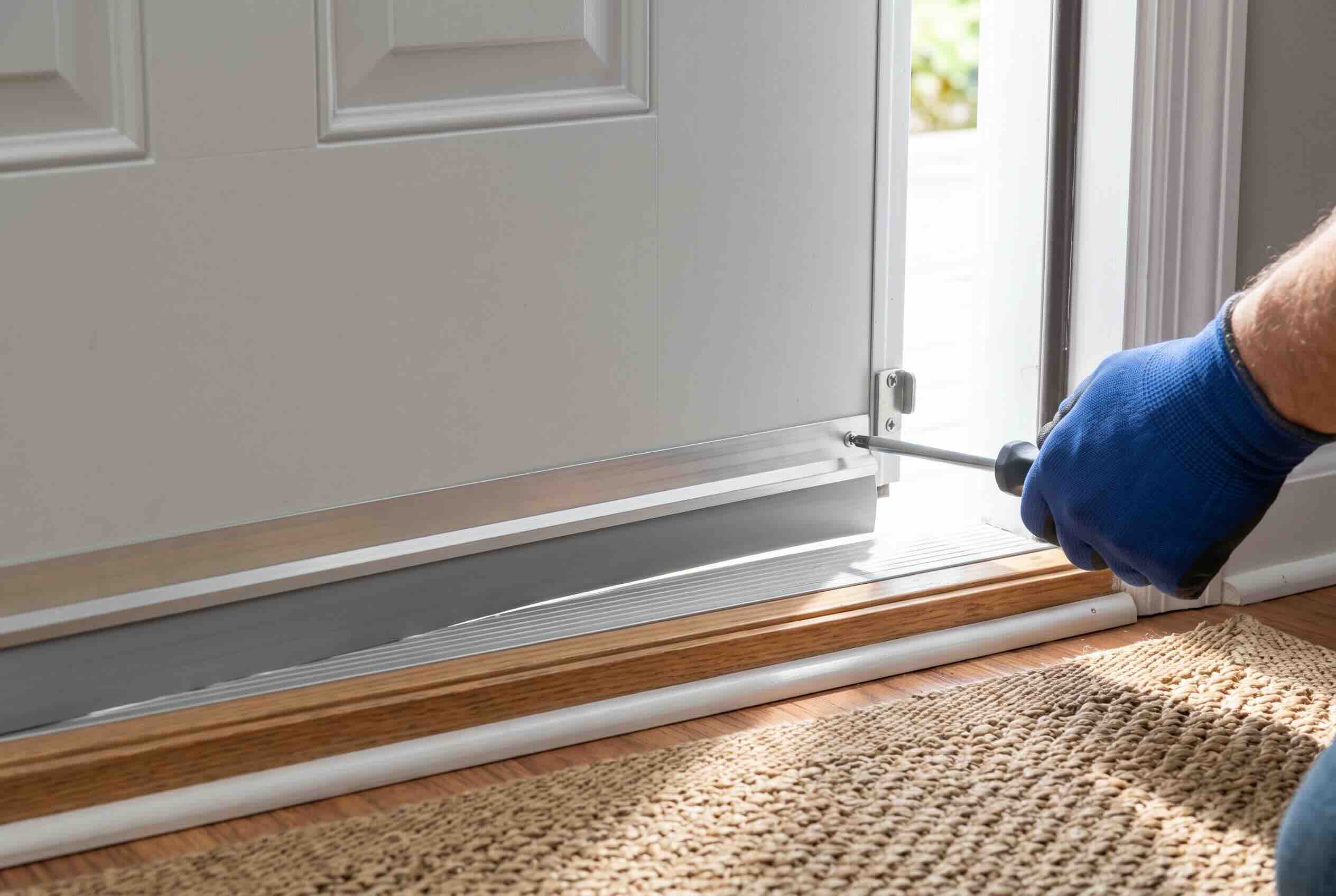 Using a screwdriver to install a new metal sweep at the bottom of a white exterior door.