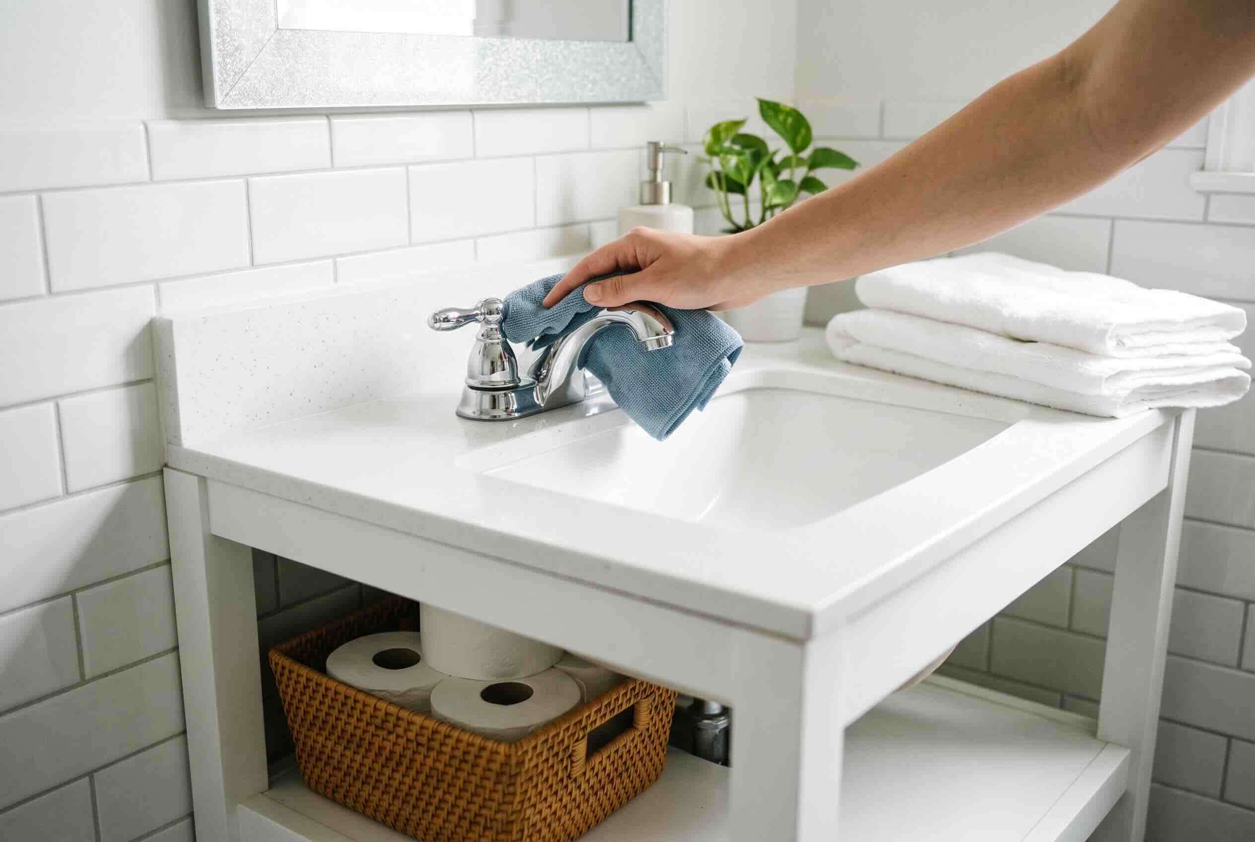A blue cleaning cloth wiping a silver faucet on a white bathroom vanity sink.