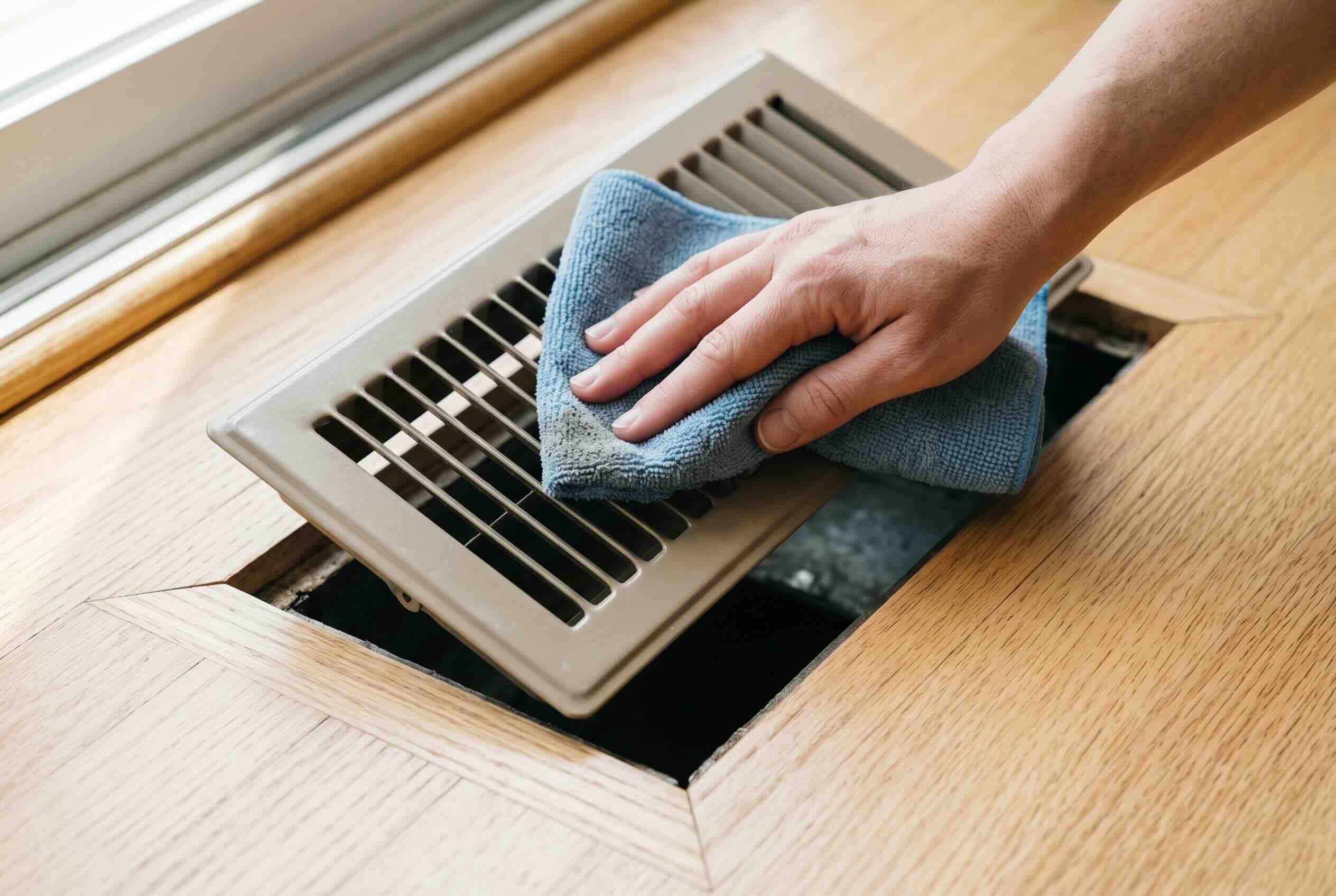 A blue microfiber cloth wiping the metal grilles of a rectangular floor air vent.