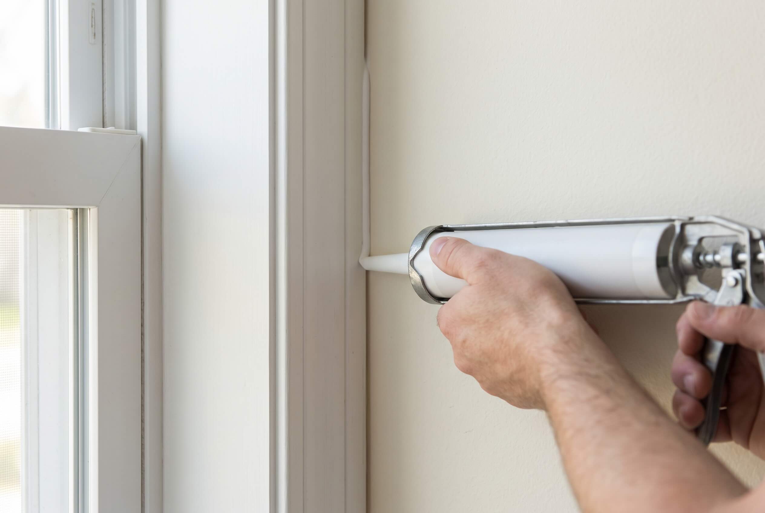 A caulking gun applying a bead of silicone caulk along the gap between a window frame and a wall.