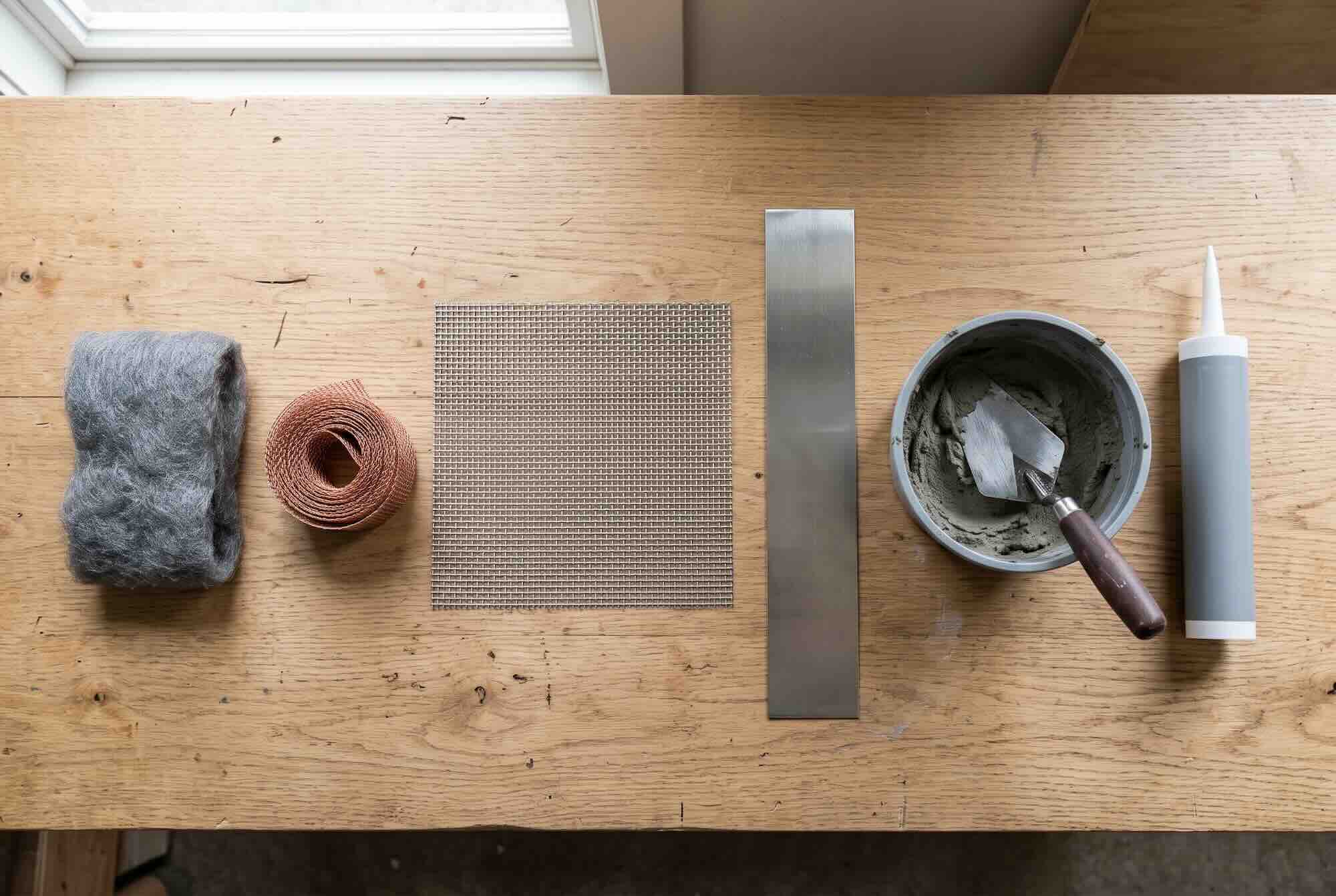 A flat lay of rodent-sealing materials on a wooden table: steel wool, copper mesh, hardware cloth, sheet metal, a bucket of mortar, and a tube of caulk.