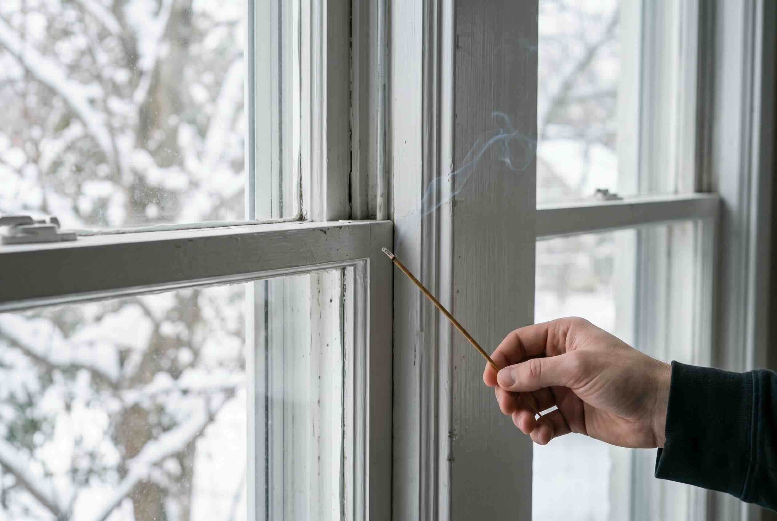 A lit incense stick releasing smoke near a white window frame to check for drafts and air leaks.