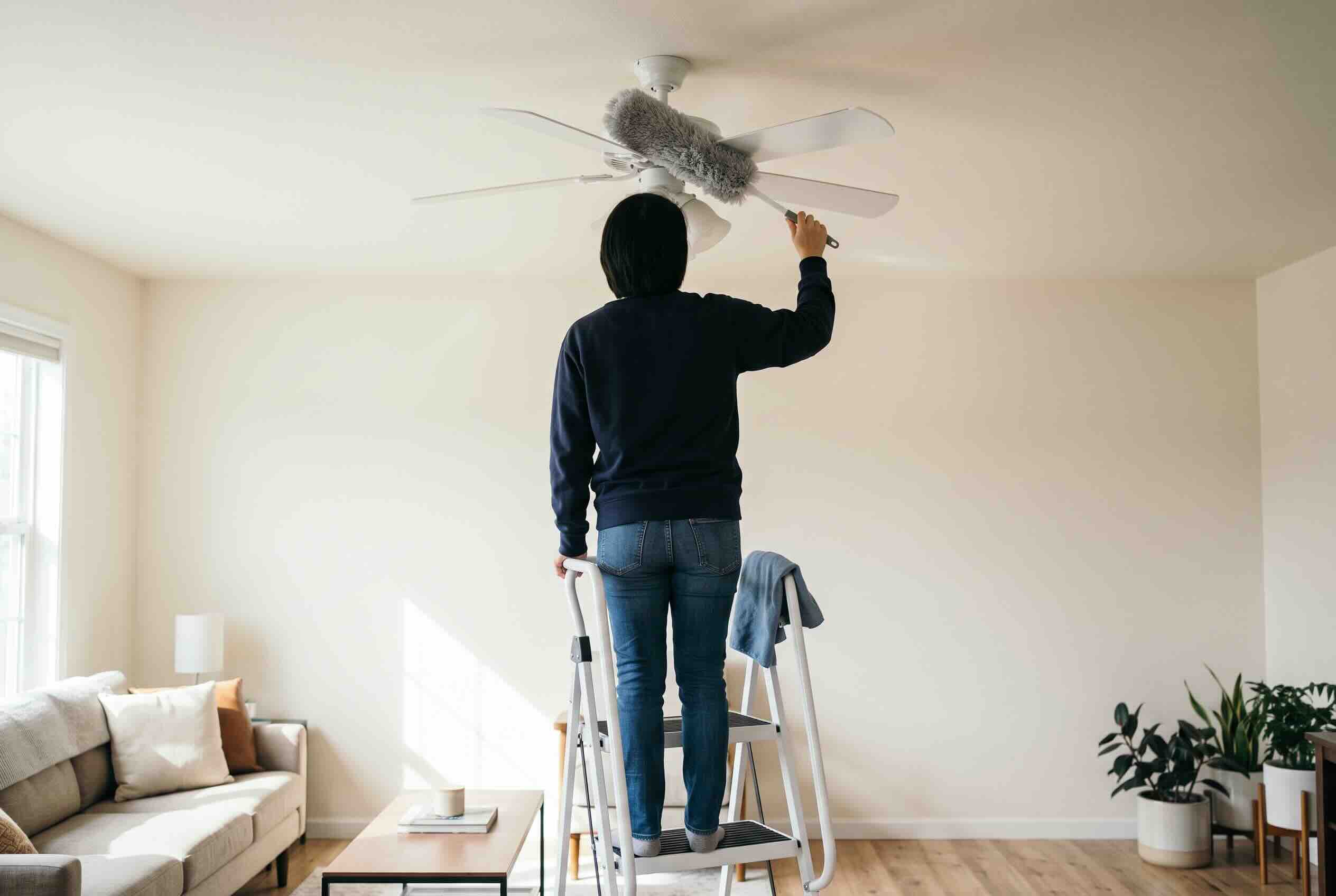 A long-handled fluffy duster reaching up to clean the white blades of a ceiling fan.