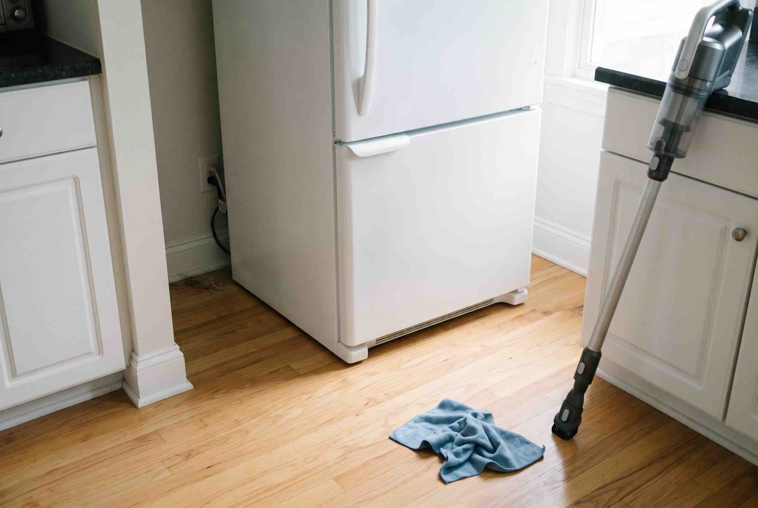 A stick vacuum and a blue cleaning cloth on the wooden floor next to a white refrigerator.