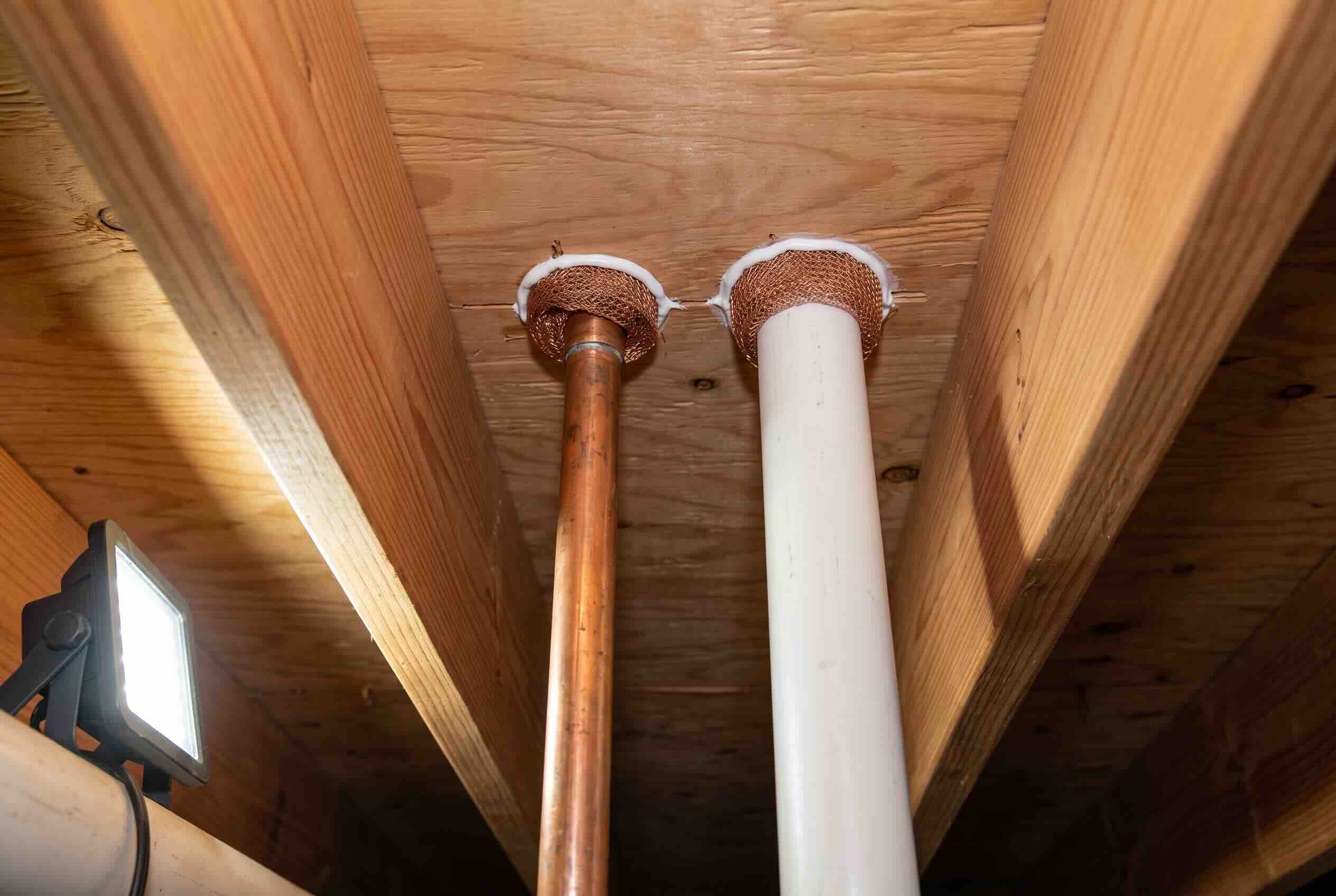 Copper mesh packed around pipes entering a wooden floor, sealed with white caulk.