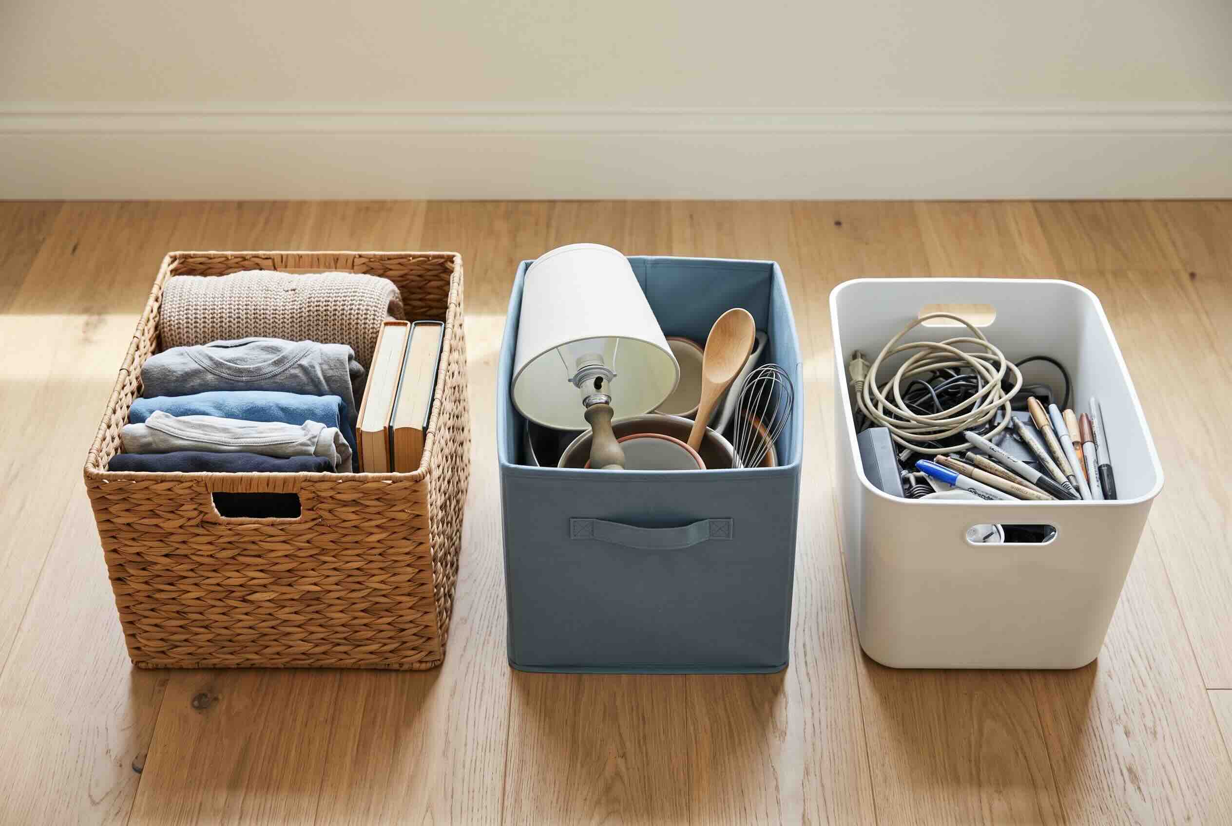 Three sorting bins on a wooden floor: a woven basket for folded clothes, a blue bin for household items, and a white bin for small electronics and pens.